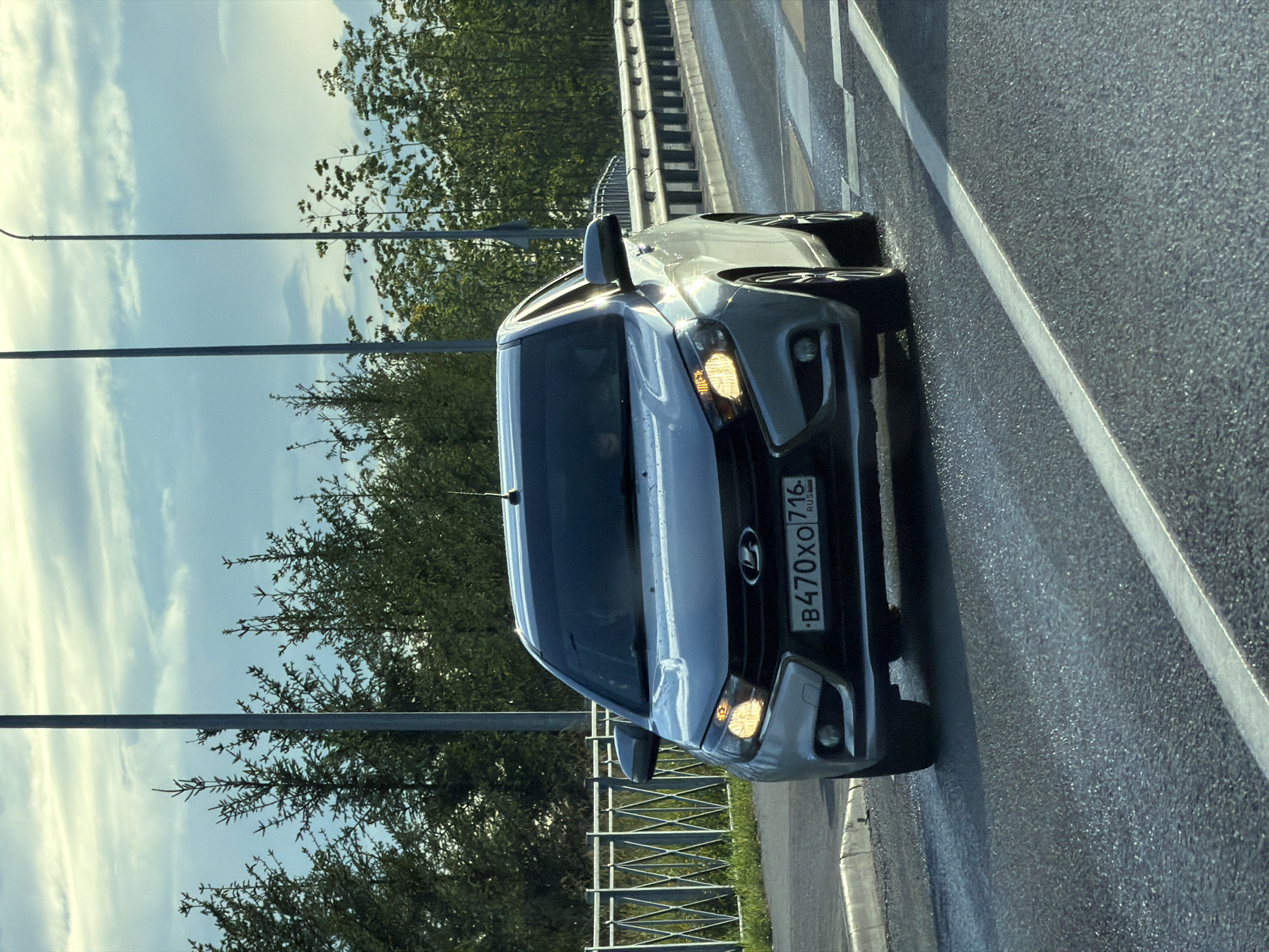 A silver Lada sedan is driving on a wet asphalt road. The car's headlights are on. The license plate reads "B470XO 716 RUS". The car is on a bridge, with a metal fence on the left and trees and lampposts in the background. The sky is cloudy. The image appears to have been taken from another vehicle. There are no visible people or interactions. The scene is likely in Kazan, Russia, given the license plate format.