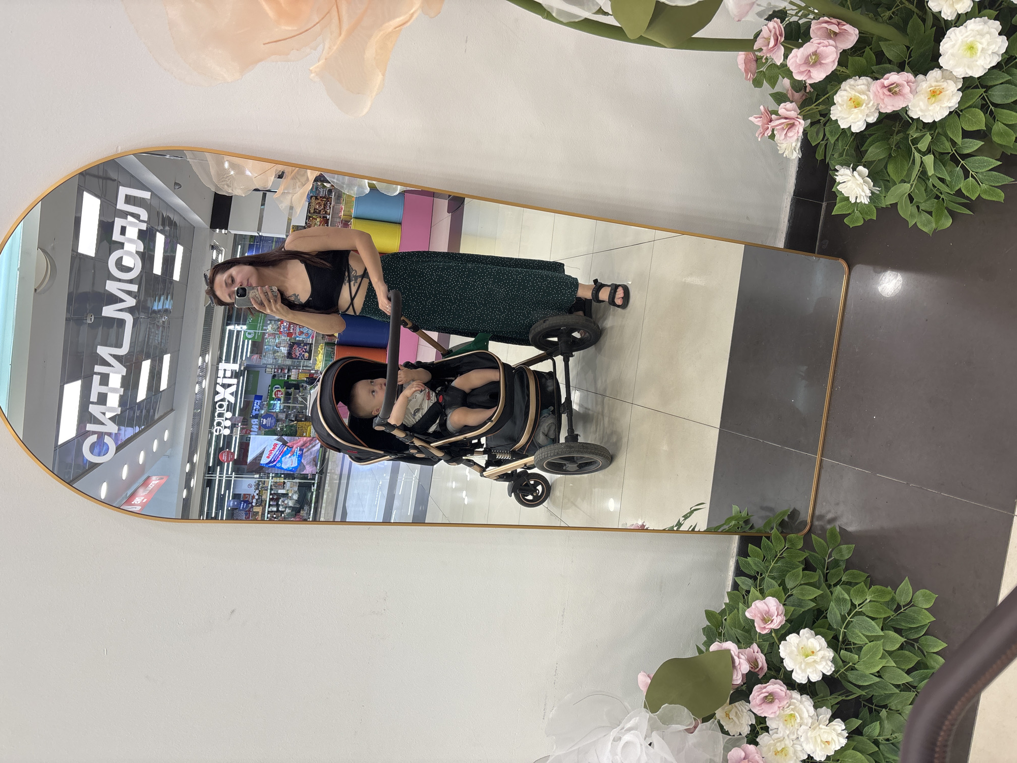 A woman is taking a mirror selfie in a mall with her baby in a stroller. The woman is wearing a black crop top and a long green skirt with white polka dots. The baby is sitting in a black stroller with gold accents. The mirror reflects the mall environment, including stores and advertisements. There are also decorative flowers in the foreground. The location is the "City Mall" in Naberezhnye Chelny, Russia, as indicated by the text reflected in the mirror.
