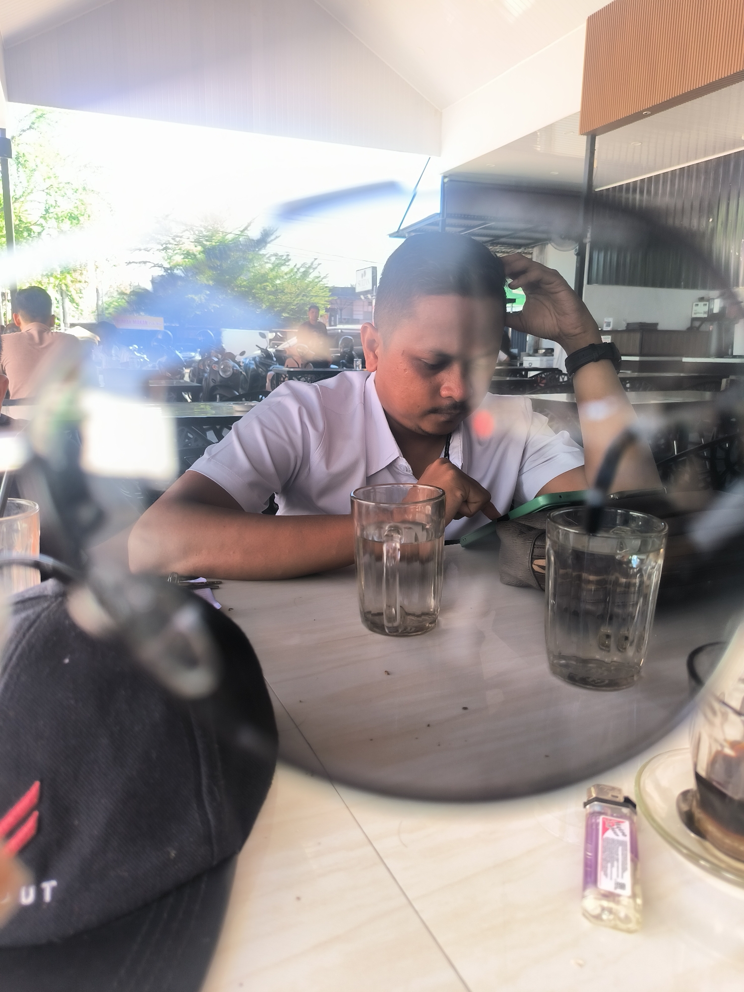 A man sits at an outdoor table in Lhokseumawe, Indonesia, wearing a short-sleeved white shirt and a black watch. He rests his head on his left hand and looks at a green-cased mobile phone on the table. Two glasses of water are on the table in front of him. A black baseball cap with red embroidery, a lighter, and a cup of coffee are also visible. The scene is viewed through the reflection of sunglasses or a lens. In the background, other patrons, motorbikes, and buildings are visible under a bright, sunny sky.