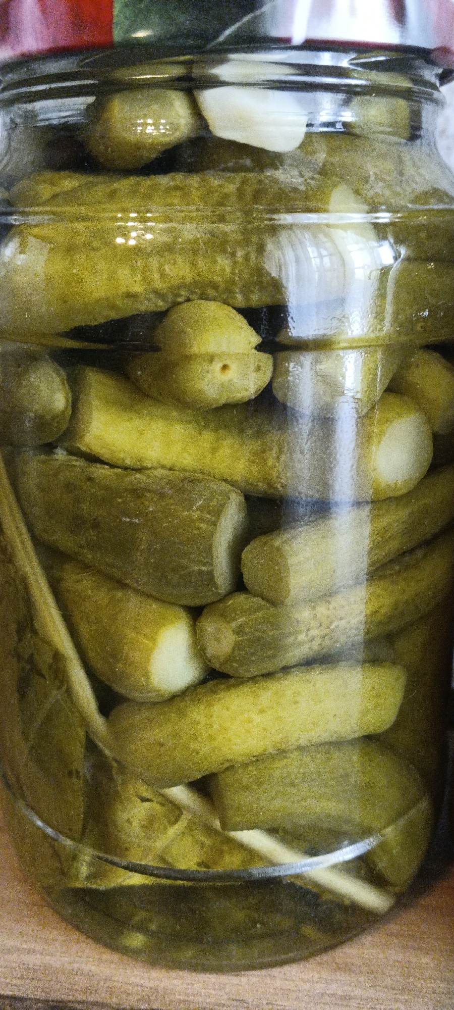 A close-up, vertically oriented, eye-level, medium shot captures a glass jar filled with numerous whole and cut pickles. The pickles are a light green color and appear to be preserved in brine. The jar is packed tightly with the pickles, some of which are cut into shorter pieces. The texture of the pickles is bumpy and rough. Reflections of light are visible on the surface of the glass jar, creating bright highlights and obscuring some of the pickles within. The background is a blur of muted colors, suggesting a simple setting. No specific landmarks or environmental details are discernible in the image to indicate a location in Tashkent, Uzbekistan. The image focuses solely on the contents of the jar.