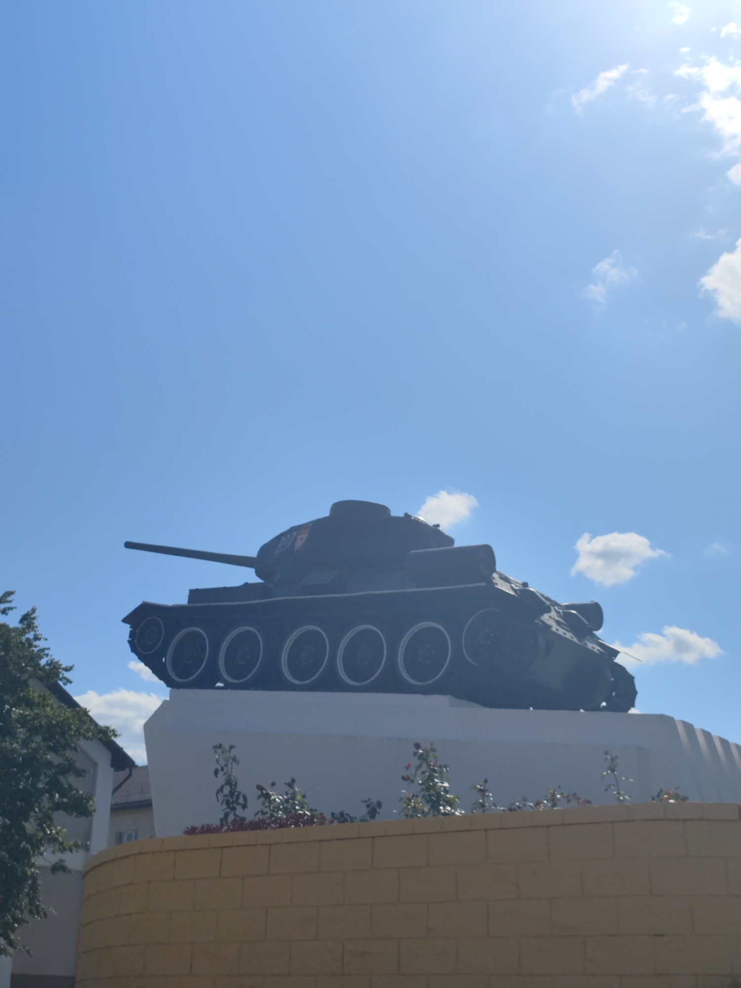 A dark-colored tank monument, identified as a T-34 tank, is positioned on a white pedestal. The tank is facing right and its barrel is extended forward. The pedestal is situated behind a yellow brick wall and in front of a building. Green trees are visible on the left side of the image. The sky is blue with scattered white clouds, and the sun is shining. This monument is located in the city of Slonim, Belarus, specifically at the intersection of Sovetskaya Street and Krasnoarmeyskaya Street.