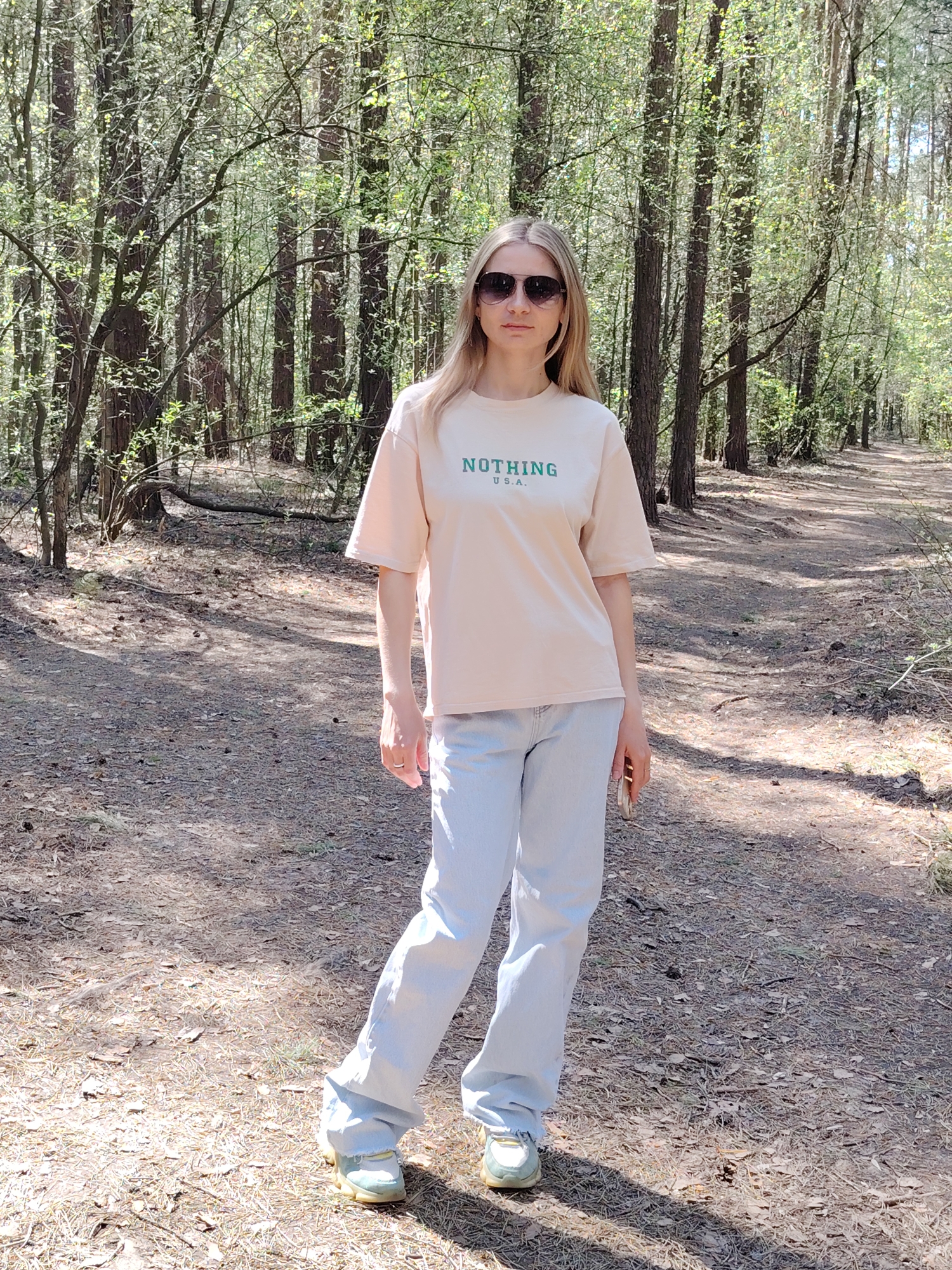 A young, fair-skinned woman stands in a forest. She wears sunglasses, a beige T-shirt with the green inscription "NOTHING U.S.A.", light blue jeans, and sneakers. The location is in a forest with tall trees and a dirt path. The forest is likely in Chaikovsky, Russia. The woman is standing and facing forward.