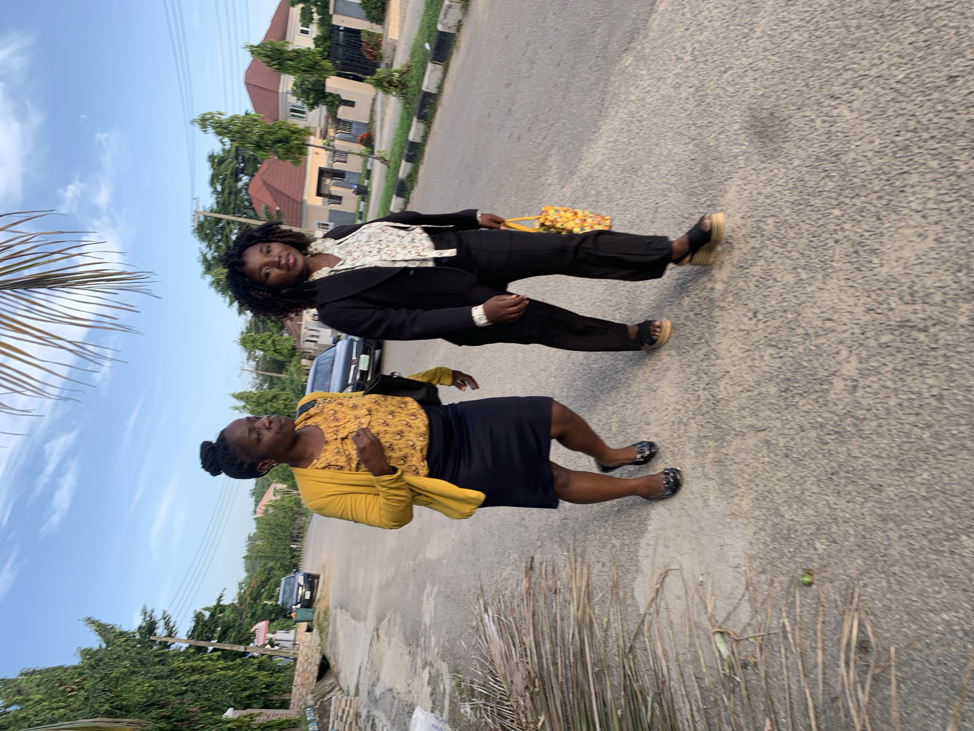 Two women are walking on a paved road in a residential area of Abuja, Nigeria, during what appears to be late afternoon or early evening. The woman on the left is wearing a yellow cardigan over a patterned yellow top and a dark blue skirt, with dark, flat shoes. She has her hair pulled up in a bun and is carrying a black bag on her right shoulder. The woman on the right is wearing a black suit jacket over a white floral blouse and black trousers, with wedge sandals. She is carrying a colorful, patterned tote bag in her right hand. Both women are facing forward and appear to be walking towards the viewer. The road is lined with trees, houses with red tiled roofs, and a sidewalk. There are parked cars visible in the background. The sky is blue with some clouds.
