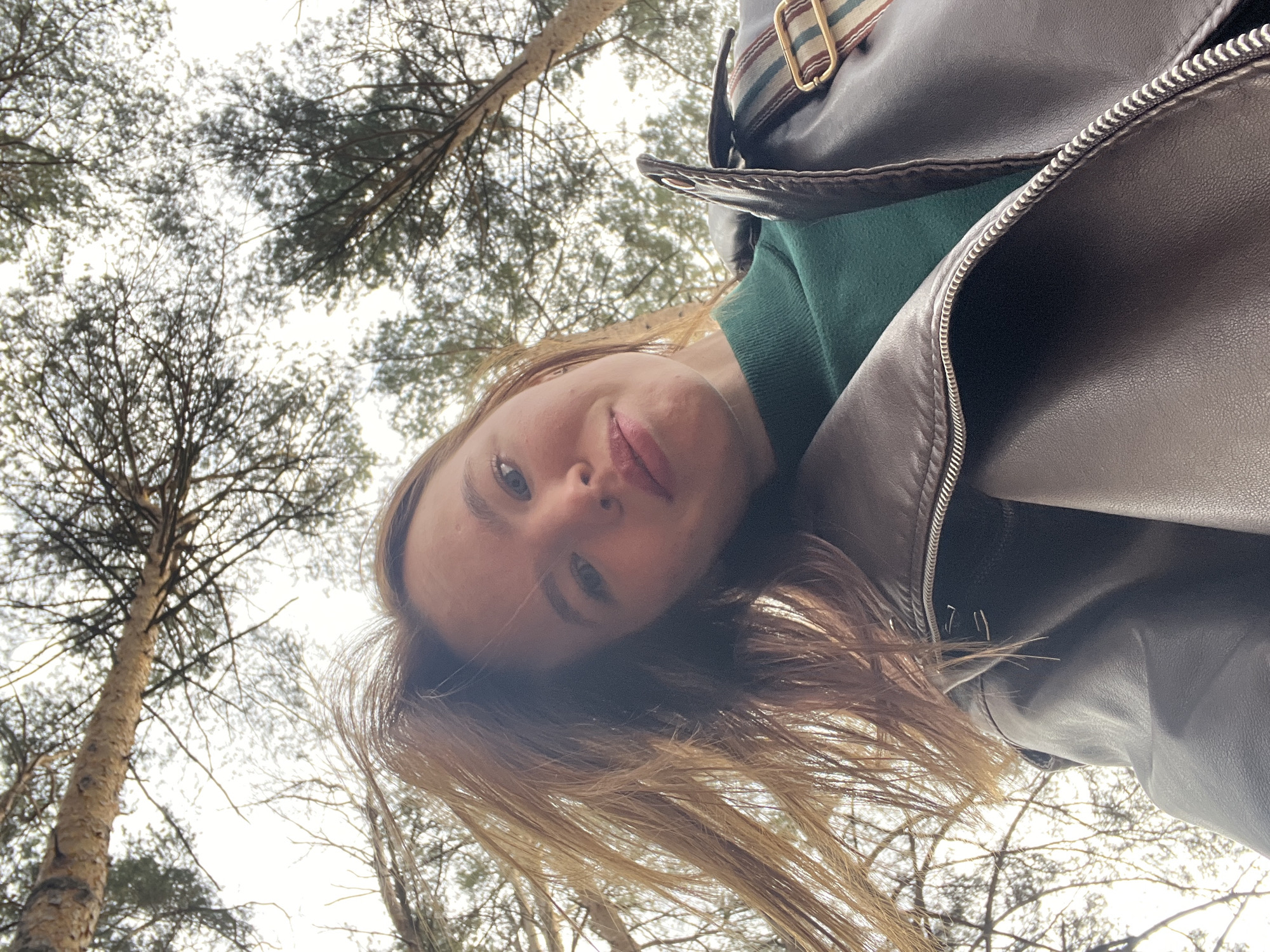 A woman with light skin and blonde hair is pictured in a low-angle selfie, with trees visible in the background. She's wearing a green sweater and a dark brown leather jacket. The setting appears to be a forest with bare trees, possibly within the city of Chaikovsky, Russia. No specific actions or interactions are visible beyond the act of taking the selfie.