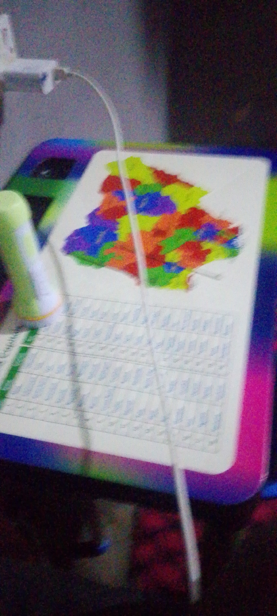 A close-up, grainy, slightly blurry image captures a colored drawing on a white piece of paper laid on a keyboard. The drawing is a map of an area with distinct regions colored in various bright hues like yellow, green, blue, purple, and orange. A thin white cord runs diagonally across the drawing. To the left of the drawing, a blurry light green cylindrical object with a white cap is visible. The keyboard appears to have rows of blue-tinged keys or markings. The background is dark and out of focus, with hints of purple and pink from the edge of what appears to be a device or surface. The lighting is dim. The image is static and does not depict any movement or action. The location is not discernible from the image.