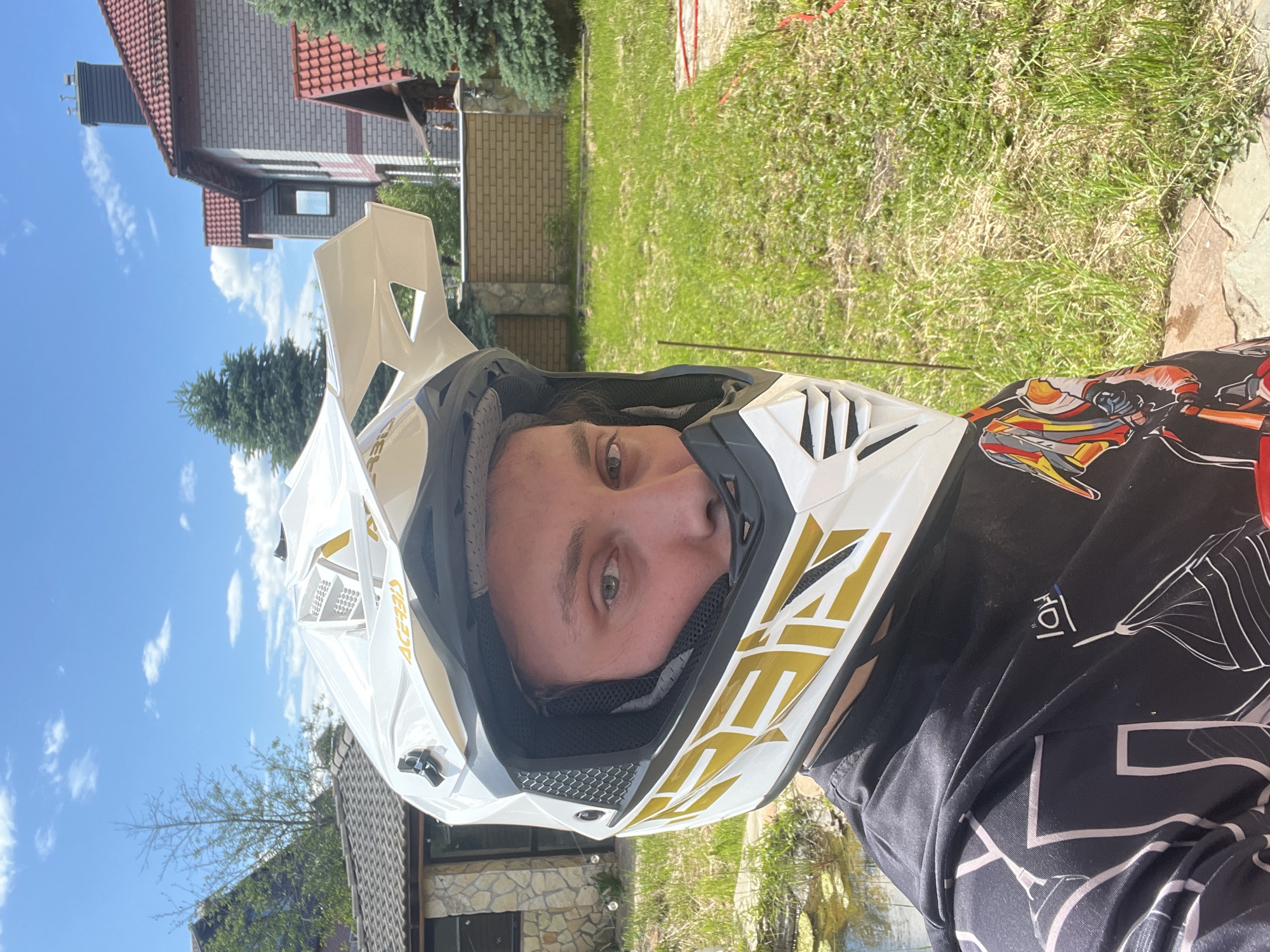 A young adult male, facing forward, is captured in a close-up outdoor portrait in Ореховка, Russia, on a sunny day with a clear blue sky and scattered white clouds. The subject wears a white motocross helmet with gold and black "ACERBIS" branding, obscuring most of his face except for his eyes and the lower portion of his nose. He is clad in a black long-sleeved jersey featuring intricate white, red, orange, and yellow graphic designs. To the left, a small pond with visible water plants is present, bordered by a building with a stone-clad base and a dark tiled roof, and a sparse, light-barked tree. To the right, a two-story house with light-colored brick walls, a red tiled roof, and a dark chimney is visible behind a beige brick fence with stone pillars. Evergreen trees are interspersed throughout the background. The ground in the foreground is covered in green grass with some dry patches and weeds, and a red cable or hose lies on the far right.
