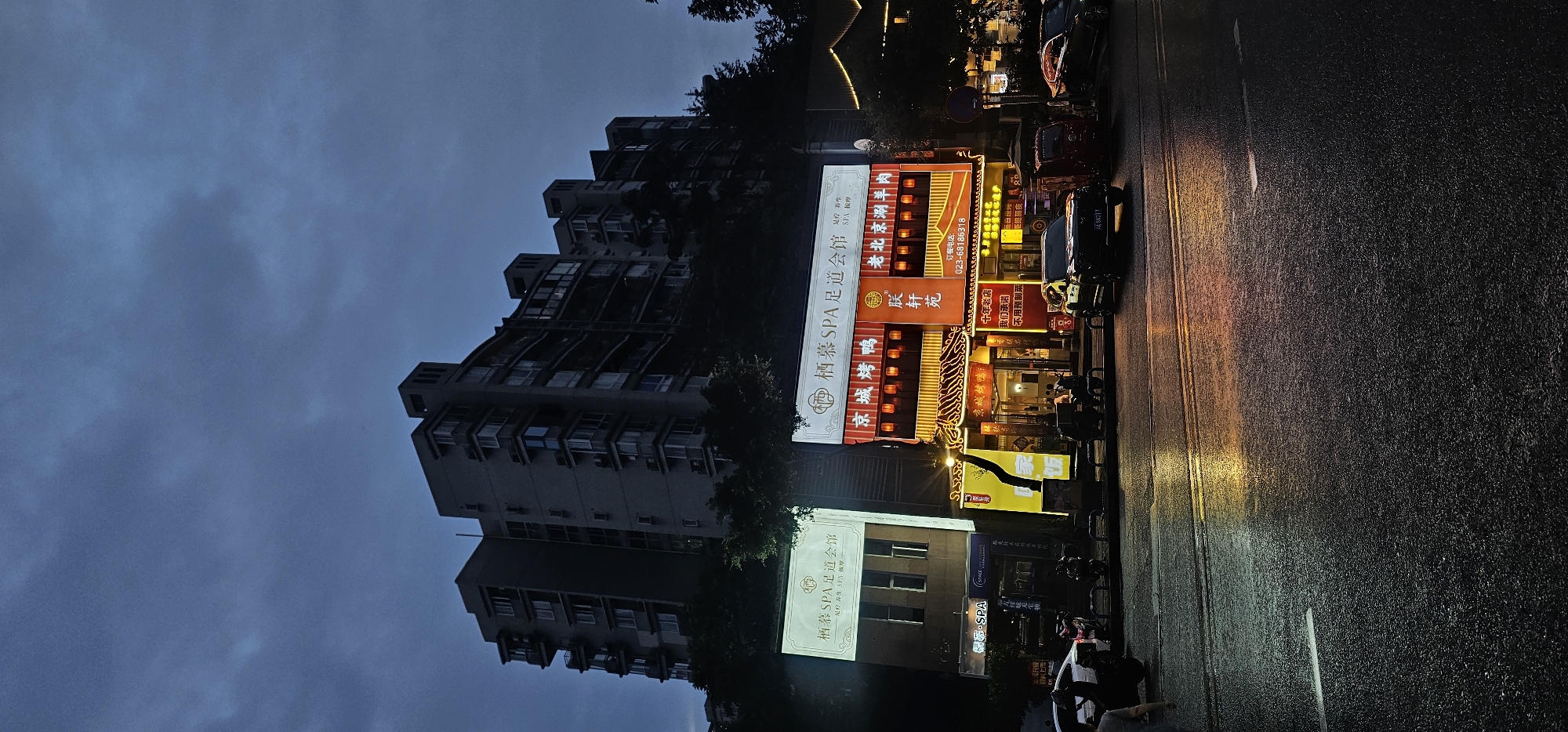 The image depicts a street scene at dusk or night in Jiulongpo District, China, with a large, modern apartment building towering over commercial establishments. The sky is dark and overcast. In the foreground, a wet asphalt road reflects the neon lights of the businesses. Several vehicles, including cars and motorcycles, are parked along the roadside. One black SUV is prominently featured in the center of the image.
The commercial buildings are illuminated with bright, colorful signage. The building on the right has a large, orange and red sign with Chinese characters, advertising "老北京涮羊肉" (Old Beijing Hot Pot) and "京城烤鸭" (Beijing Roast Duck). There are also lanterns visible along the facade, suggesting a traditional Chinese restaurant. The building to the left has a lighter colored sign advertising "SPA".
The wet road surface indicates recent rain, creating reflections of the lights. The overall atmosphere is one of an urban environment at night, with a blend of residential and commercial activity. The lighting suggests it is after sunset, and the darkness of the sky indicates the time of day.