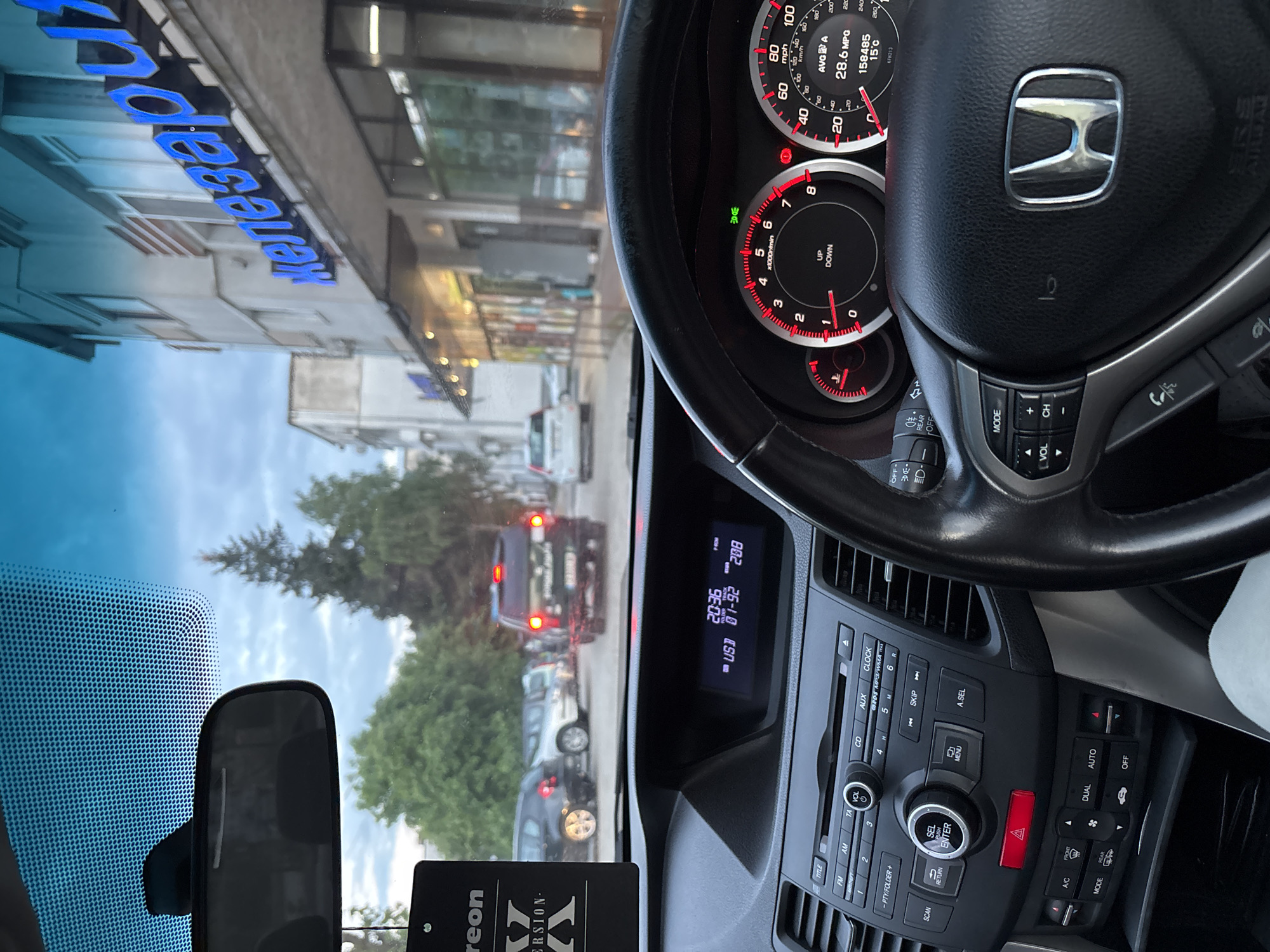 The image shows the interior of a Honda car, specifically the dashboard and steering wheel. The instrument cluster is visible, displaying speed and RPM. The car stereo system is also in view, showing a time of 20:36 and a date. A car air freshener hangs from the rearview mirror. Outside the windshield, the reflection shows a street scene with trees, buildings, and traffic. The reflection indicates a sign in blue letters that reads "Железари" (Zhelezari). Based on the visible sign and the location's name, this image was captured in Zlatograd, Bulgaria. The car is stopped in traffic.