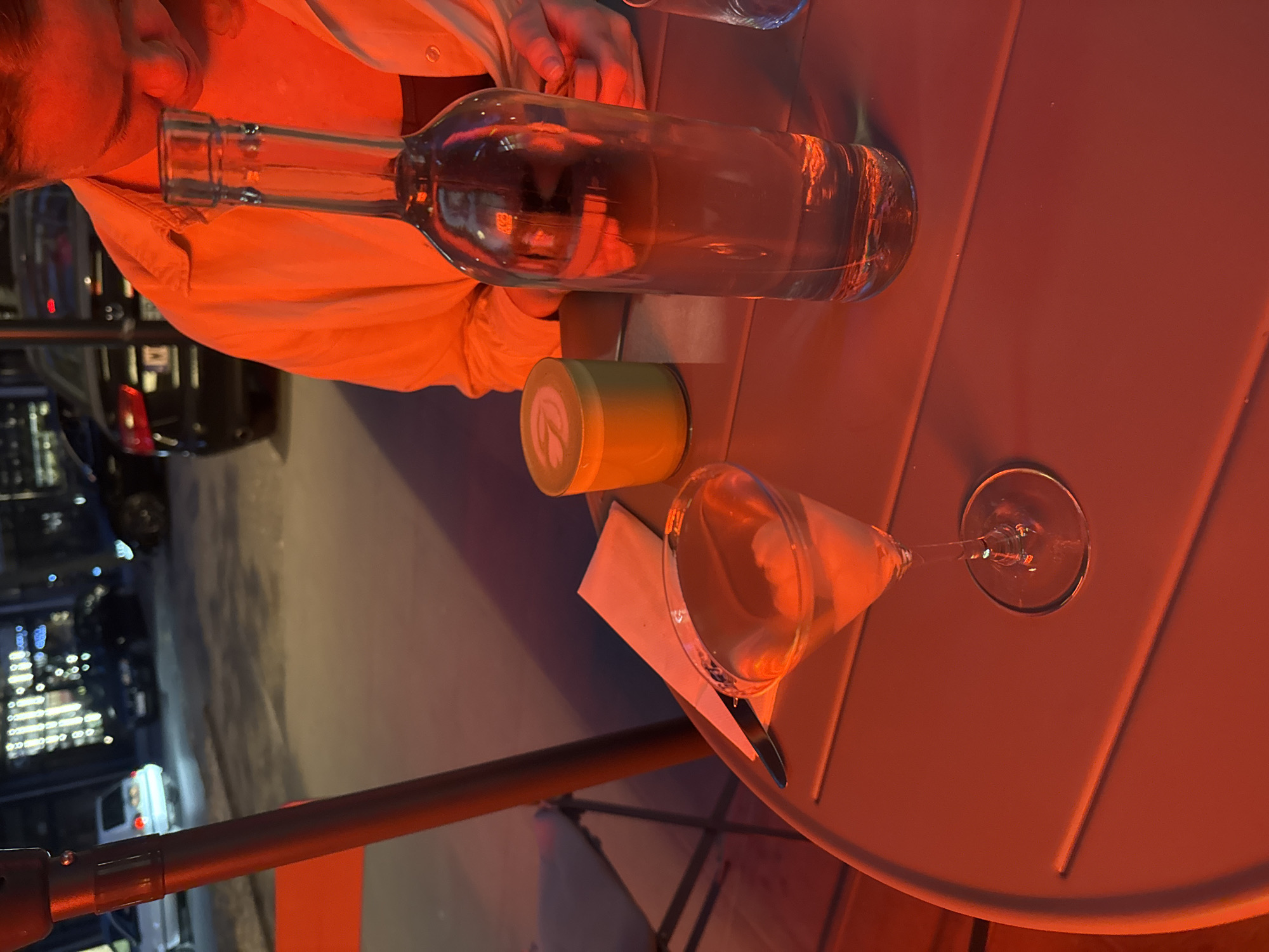 The image shows a table at an outdoor cafe, likely in the evening. A person is partially visible in the upper left, wearing a light-colored shirt. On the table, there is a glass bottle filled with a clear liquid, a small green container, a napkin with a fork on it, and an empty martini glass. The table is made of orange metal and has a ribbed surface. Outside the cafe area is a street scene showing the city of Warsaw, Poland. There are buildings with lit windows and cars driving on the street. The overall color cast of the image is orange.