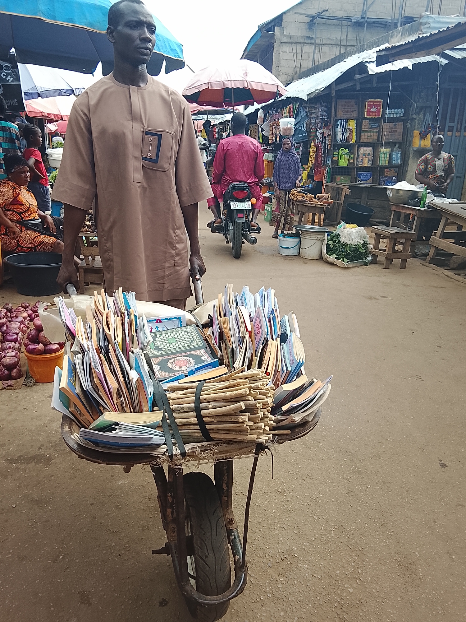 A full-shot, eye-level, outdoor, daytime photograph shows a man in a light brown dashiki pushing a wheelbarrow filled with books in a marketplace in Akwanga, Nigeria. The man, who is the central figure, is facing slightly to the right and looking forward. He has dark skin and short, dark hair. The wheelbarrow is also in the foreground, with its front wheel visible. The books are neatly stacked in the wheelbarrow, some are upright and others are lying flat. They are of various sizes and colors.
In the background, the marketplace is bustling with activity. There are stalls with goods displayed, including food items and other merchandise. Several people are visible in the background, some are walking, others are standing. A woman in an orange and brown patterned dress is seated on the left, and a young boy in a red shirt is standing behind her. A motorcycle with a rider in a pink outfit is moving away from the camera. The environment appears to be a typical African marketplace, with corrugated metal roofs over some of the stalls and a dusty ground. The overall lighting suggests it is a bright, sunny day.