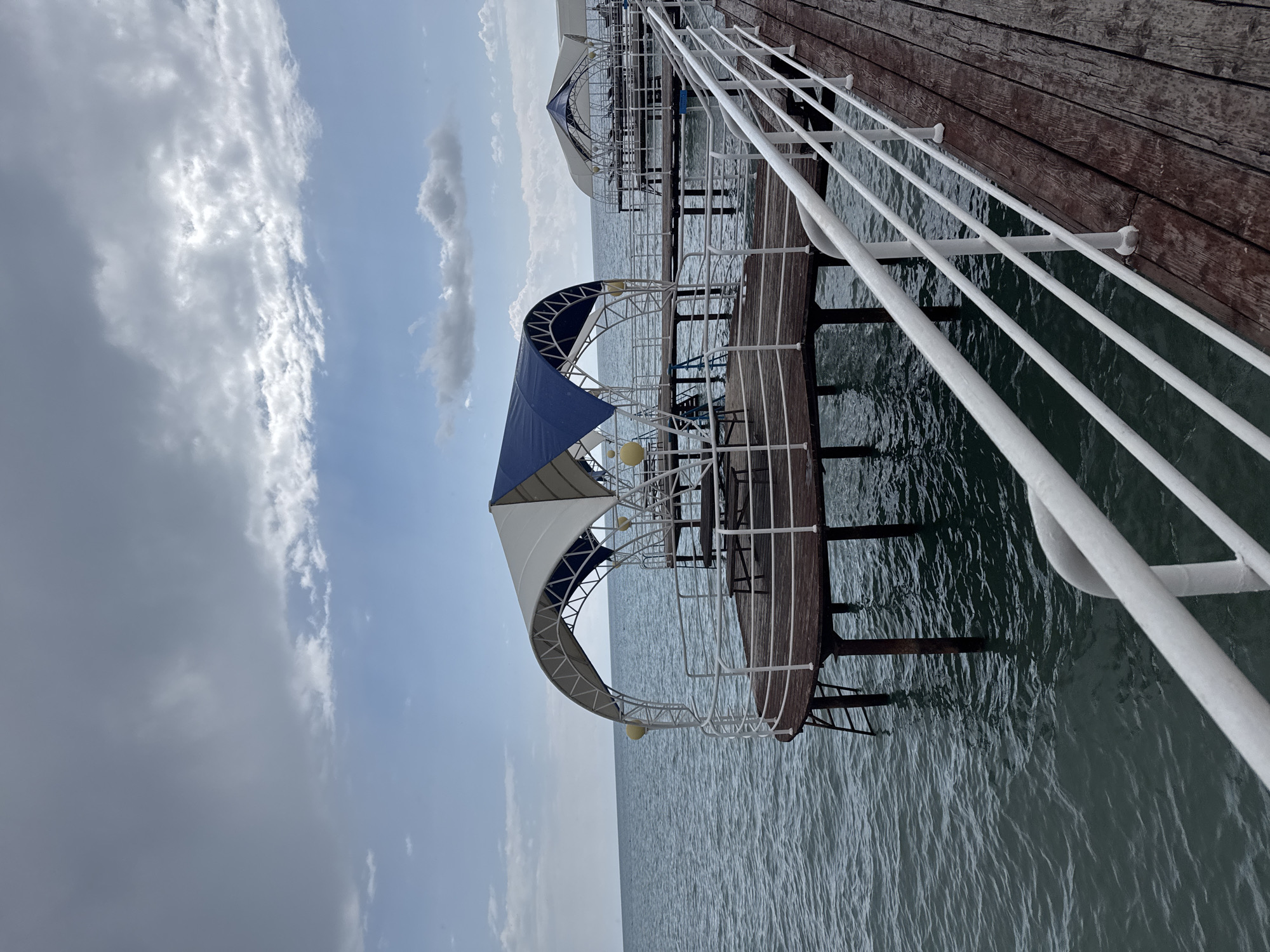 The image shows a wooden pier extending over a body of water. On the pier, there are several covered seating areas with white and blue canopies. The water is calm with some ripples. The sky is overcast with grey clouds. The pier has white railings. The scene appears to be located in a resort area. Sary-Oy is a village on the northern shore of Lake Issyk-Kul in Kyrgyzstan, known for its beaches and resorts. This pier is likely part of one of these resorts. There are no people visible in the image.