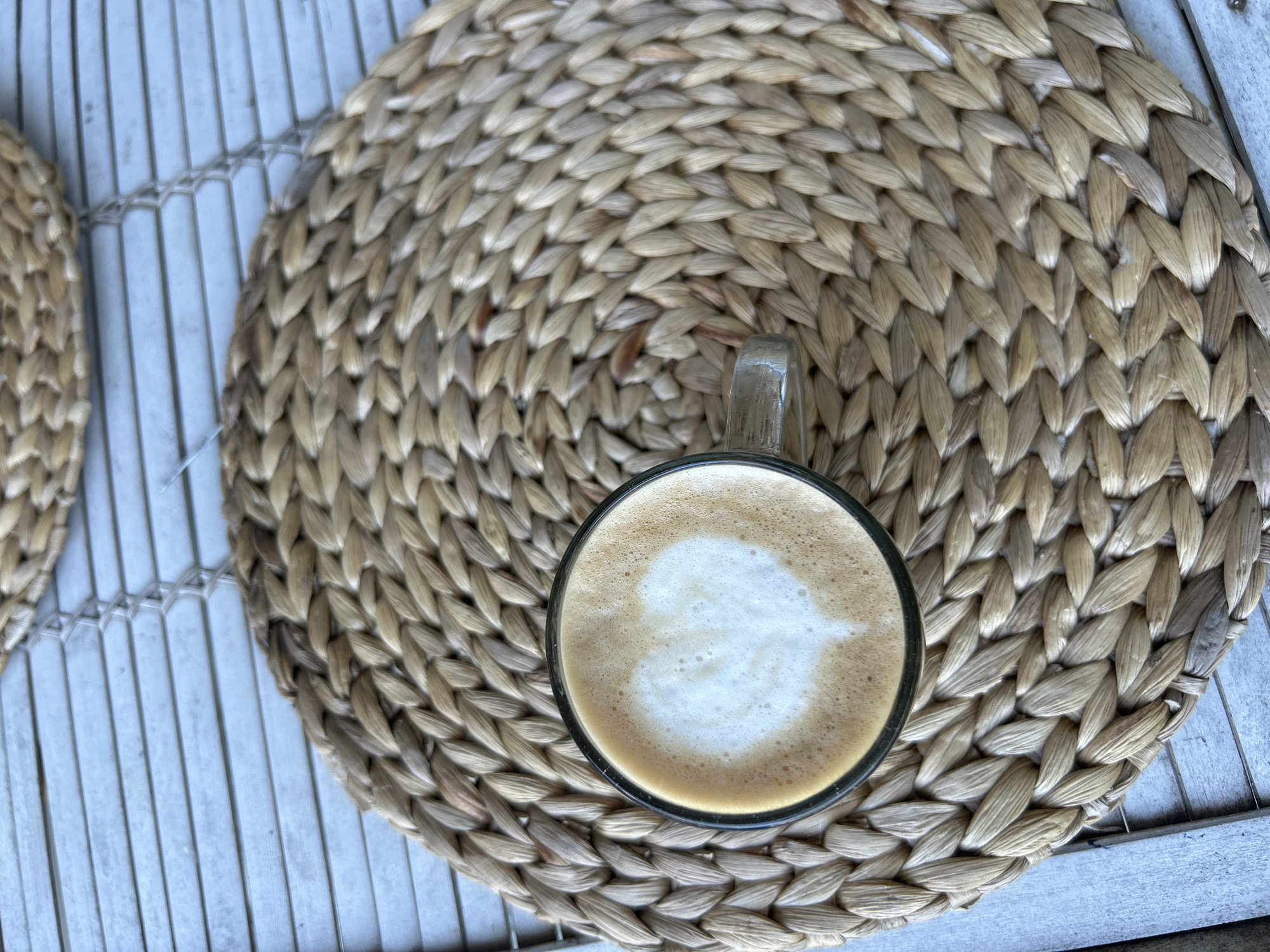 A top-down shot shows a clear glass mug filled with a cappuccino set on a woven circular placemat. The placemat is resting on a white-painted wooden slat table. The cappuccino has latte art in the center. The picture was taken in Kuta Selatan, Indonesia. There are no people present.