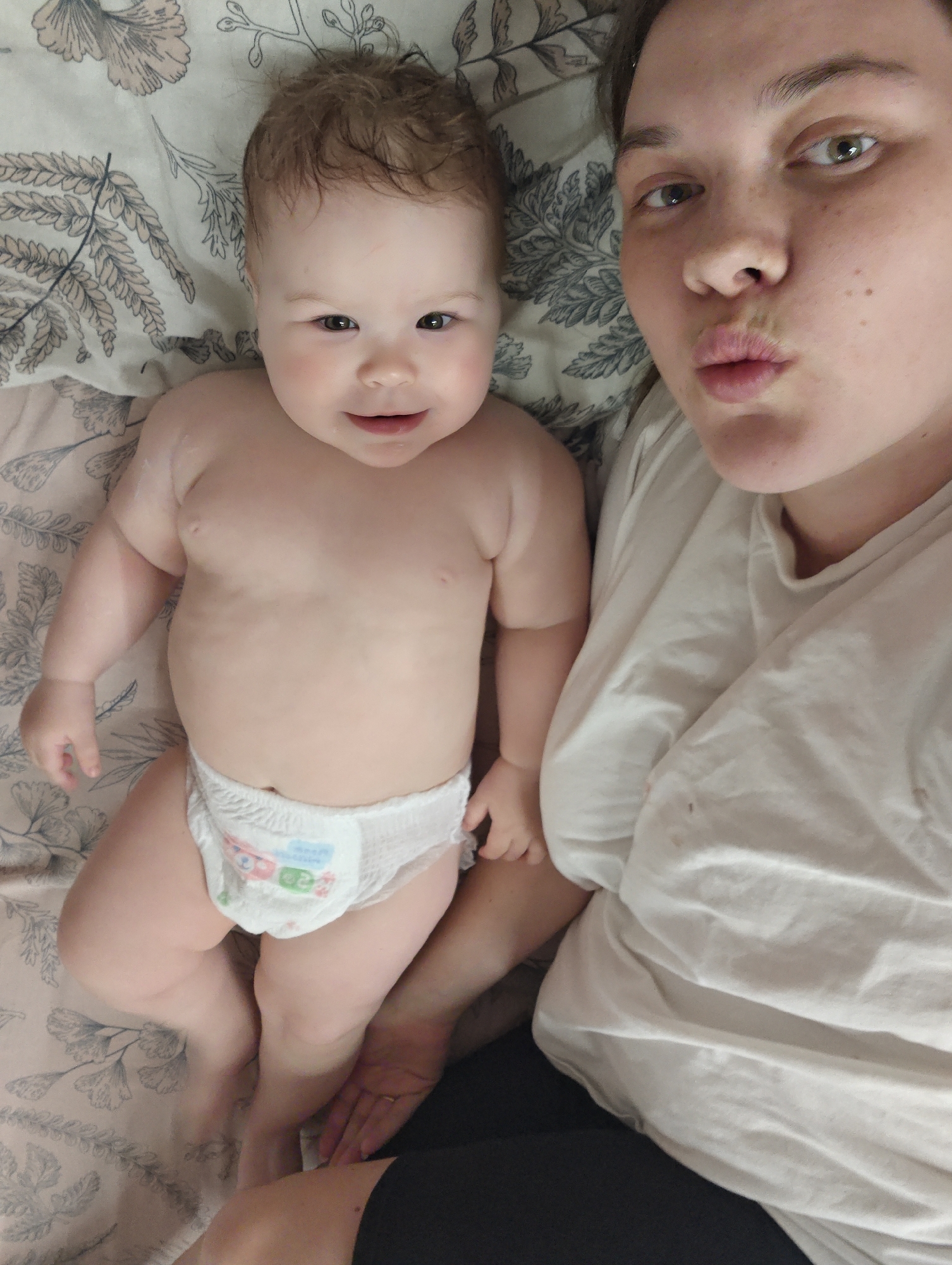 This is a close-up, high-angle selfie taken indoors in Chaikovsky, Russia, featuring a young adult woman and a baby lying side-by-side, likely on a bed. The background is a patterned sheet or blanket adorned with a subtle floral and leafy design in muted tones of beige, grey, and light pink.
The woman, positioned on the right side of the frame, has light skin, dark eyes, and visible freckles on her face. She is looking directly at the camera with a playful expression, her lips pursed in a "kiss" shape. She is wearing a simple, light-colored (possibly white or off-white) t-shirt, and a dark piece of clothing is visible lower down. A gold-toned ring can be seen on her left hand.
Next to her, on the left, is a baby with light skin, dark eyes, and light brown, curly hair. The baby is shirtless, revealing a plump torso, and wears a white disposable pull-up diaper. The diaper features faint, colorful cartoon characters and numbers, suggesting a size indicator like '2' or '3'. The baby looks towards the camera with a wide-eyed, happy smile, conveying a sense of joy and contentment.
The image captures an intimate and affectionate moment, likely between a mother and child, characterized by their playful expressions and close proximity. The lighting is soft and even, consistent with an indoor setting.
