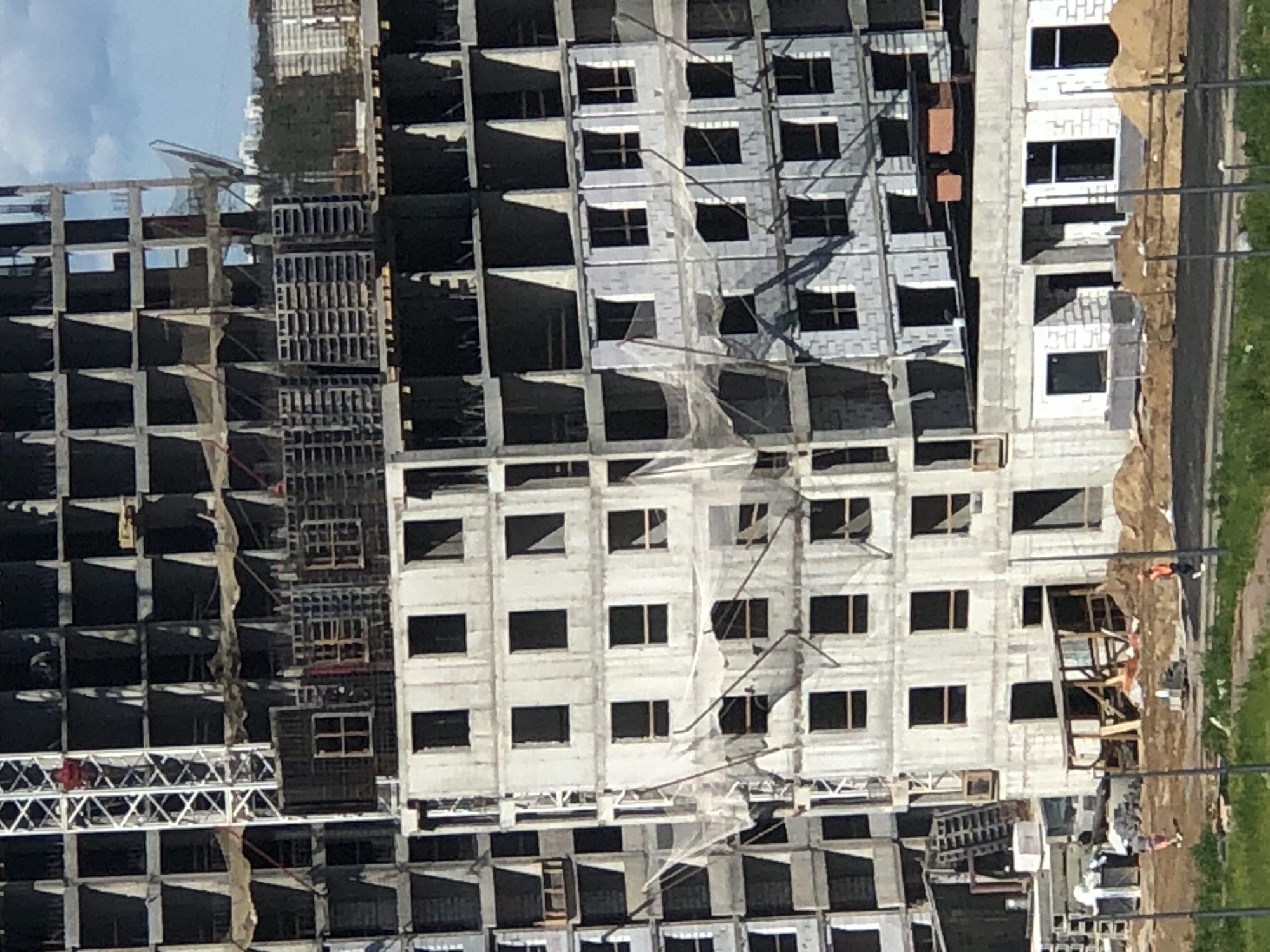 The image displays a large-scale construction site in Saint Petersburg, Russia, featuring multiple reinforced concrete high-rise buildings under various stages of construction. The structures reveal exposed frameworks of columns and floor slabs, with some sections partially enclosed by concrete block walls that define numerous rectangular window openings.
A prominent white lattice tower crane stands vertically on the left side of the primary building, with one individual in a red shirt visible within its mast structure at an elevated position. Several upper floors exhibit stacked dark grey metal or wooden formwork panels. White safety netting is draped across multiple mid-level floors on the central and right sections of the construction.
The ground level consists of disturbed earth with piles of sandy soil and scattered construction materials, including concrete blocks. Temporary wooden structures, possibly shelters or scaffolding, are also present on the ground. Three individuals are identifiable: one in the red shirt within the crane, one person in light clothing walking right on the dirt ground in the lower-left, and another person wearing an orange high-visibility vest walking right on the dirt ground in the lower-right.
The scene is illuminated by bright daylight, indicated by strong shadows cast by the buildings and the crane. The sky shows patches of blue with scattered white clouds. A strip of green grass occupies the immediate foreground, and distant urban structures along with trees are visible on the horizon.