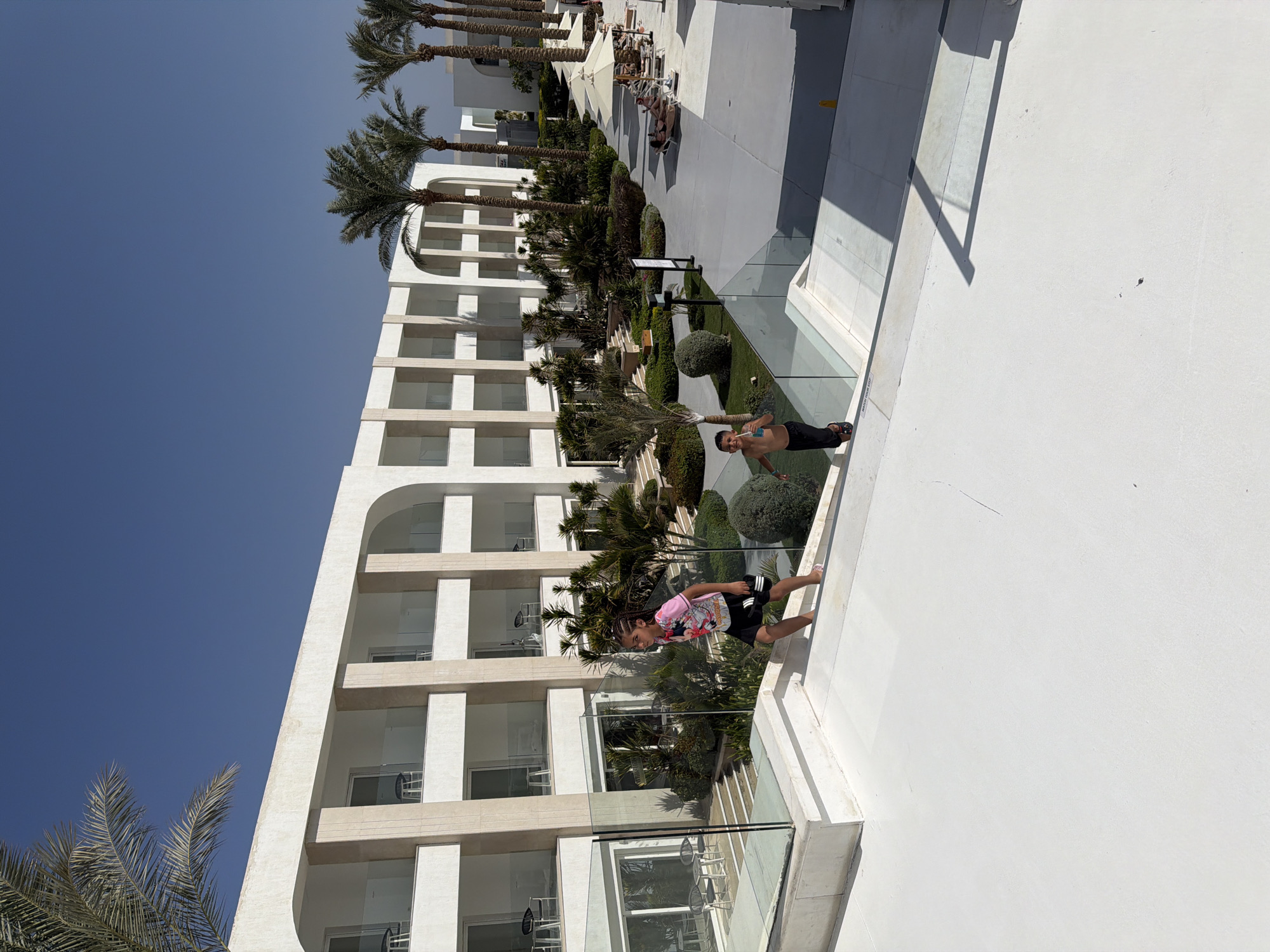 The image shows a modern, multi-story resort with white walls and arched balconies, set against a clear blue sky. Palm trees are scattered throughout the property, with a row of taller palm trees lining a walkway to the right. Two young people, one in a pink t-shirt and black shorts, the other shirtless in black shorts, are running on a white outdoor area with glass railings. In the background, there are lounge chairs and umbrellas by a pool area, where other people are seen relaxing. The resort appears to be located in Sharm El Sheikh, Egypt, known for its sunny climate and resort atmosphere.