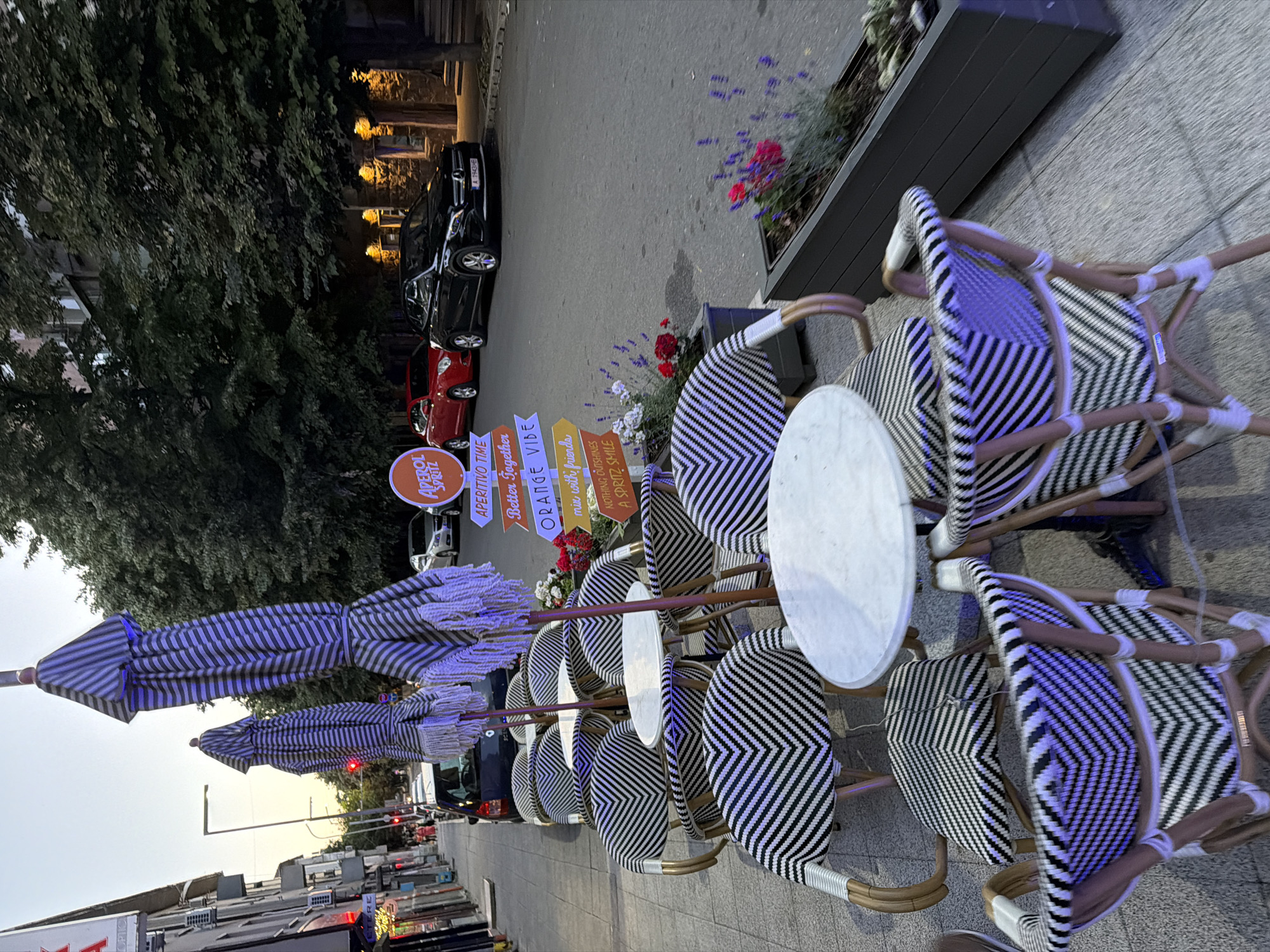 The image depicts an outdoor seating area of a cafe or restaurant located on a street in Burgas, Bulgaria. The foreground features several tables and chairs with a distinctive black and white striped wicker pattern, set up on a paved sidewalk. Two closed umbrellas, also with a striped pattern, stand next to the tables. In the background, cars are parked along the street, and buildings line the opposite side. A sign with "APEROL SPRITZ" and several directional arrows pointing to phrases like "APERITIVO TIME," "Better Together," "ORANGE VIBE," "mix with friends," and "NOTHING BUT A SPRITZ SMILE" is visible. The lighting suggests it is either dusk or early evening. There are no identifiable people in the scene.