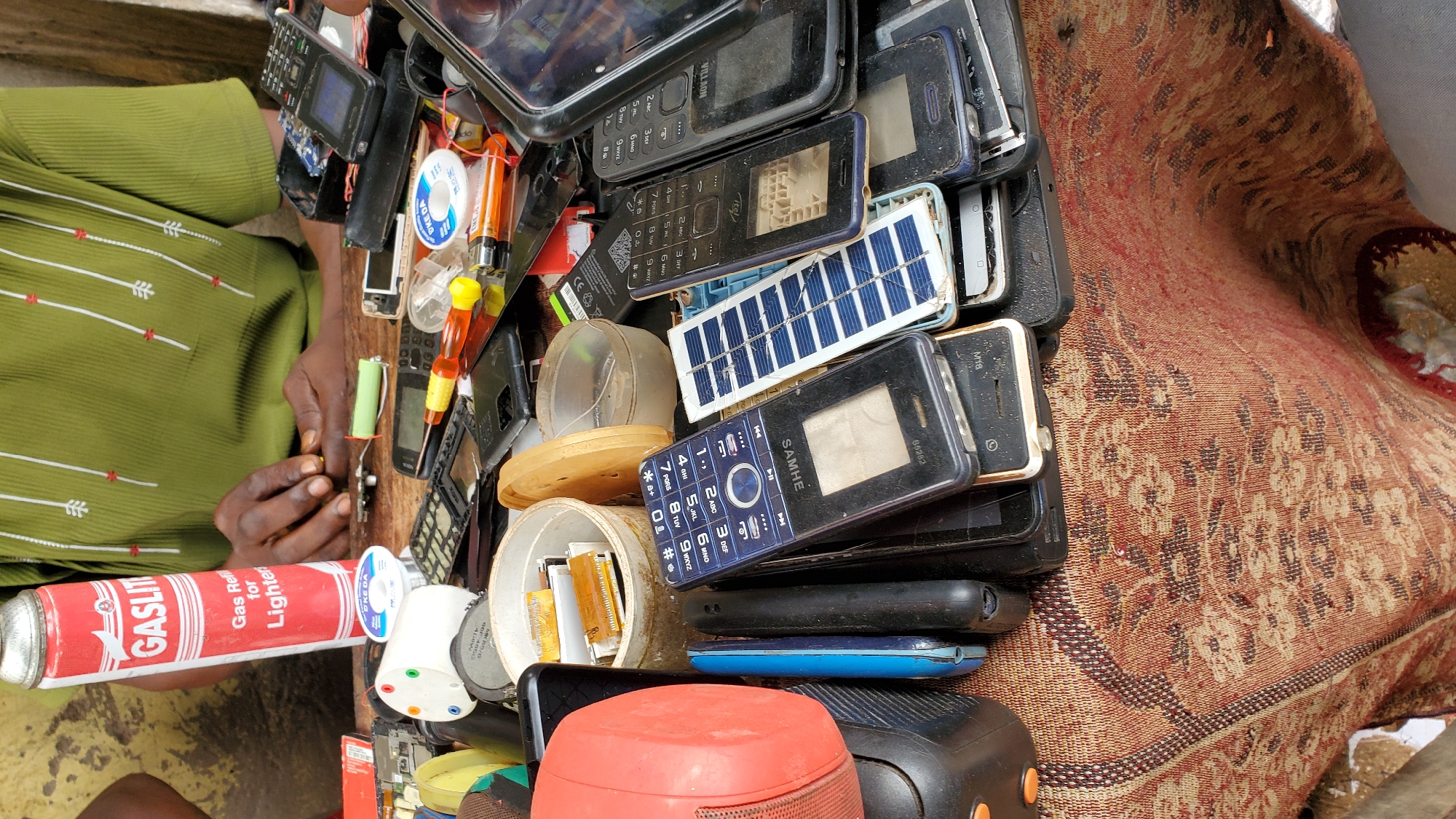 A close-up shot captures a table laden with assorted electronic components, primarily focusing on mobile phones and related accessories, suggesting a repair or recycling station. The items are densely packed, creating a cluttered yet organized workspace. Prominently featured is a red and white can of "GASLIT" lighter fluid, indicating its use in minor repairs or maintenance. Several mobile phones, in various states of disassembly or disrepair, are spread across the table. Among these, a blue and black mobile phone with a solar panel attachment stands out, hinting at alternative charging solutions or experimental modifications. Other items include batteries, wires, a soldering iron, and various small electronic parts, all contributing to the scene of an active electronics workshop. The backdrop shows the hands of an individual, likely the proprietor or technician, engaged in their work, with a green garment suggesting everyday attire. The overall ambiance points to a functional, perhaps informal, business operating within a community setting, possibly in a city like Jalingo, Nigeria, where such repair services are common.