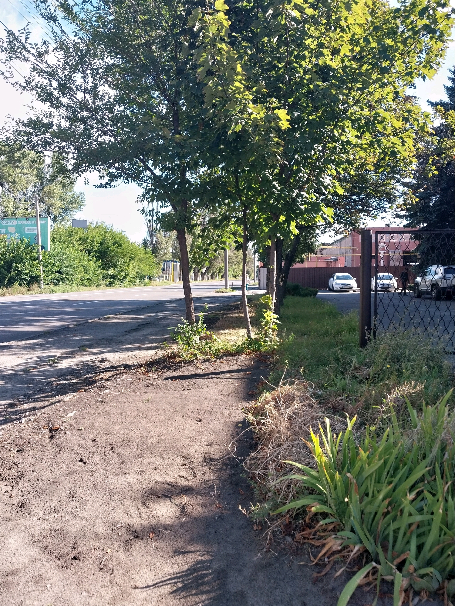 The image depicts a street scene in Dnipro, Ukraine. In the foreground, a dirt path runs alongside a paved road. Several trees with green foliage are present, casting shadows on the path. Beyond the road, there is a row of dense green bushes and then a building with a reddish-brown fence and a metal gate. Several cars are parked within the fenced area, including a white sedan and a white SUV. A person is walking near the parked cars. The sky is visible in the background, with a few clouds. The overall impression is a sunny day in a somewhat industrial or commercial part of the city, with a mix of natural and man-made elements.