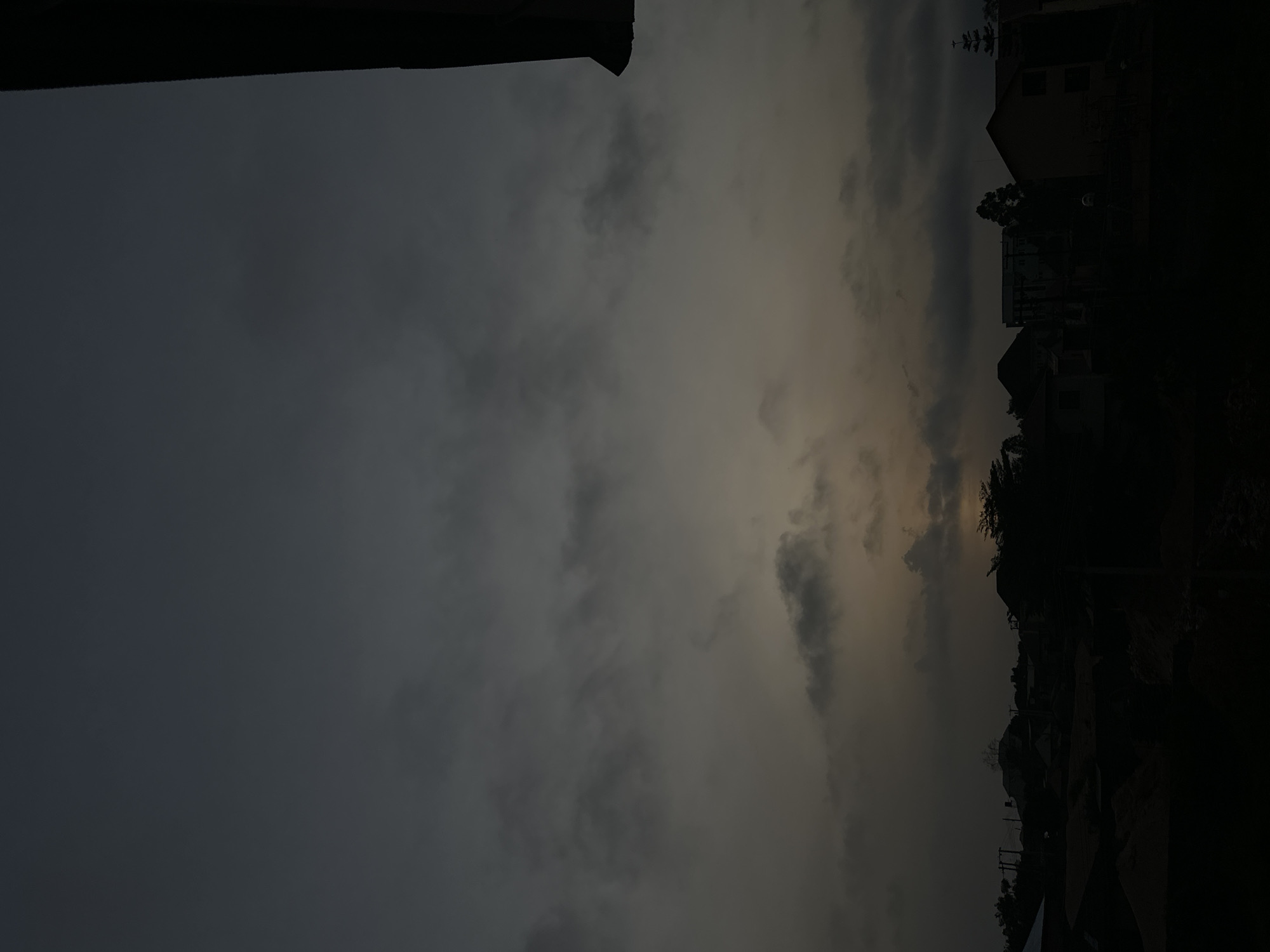 The image shows a darkened, obscured view of a neighborhood in Jos, Nigeria, under a cloudy sky. The houses are visible in silhouette, mostly to the right of the frame, with varying architectural styles. A heavy overcast sky occupies the majority of the image, creating a dim and gloomy atmosphere. There is a slight hint of lighter tones, possibly from a breaking sunset or diffused light, piercing through the clouds, but the overall visibility is low. A structure, possibly a building overhang or a portion of a roof, is visible in the upper left corner. No people are visible in the scene.