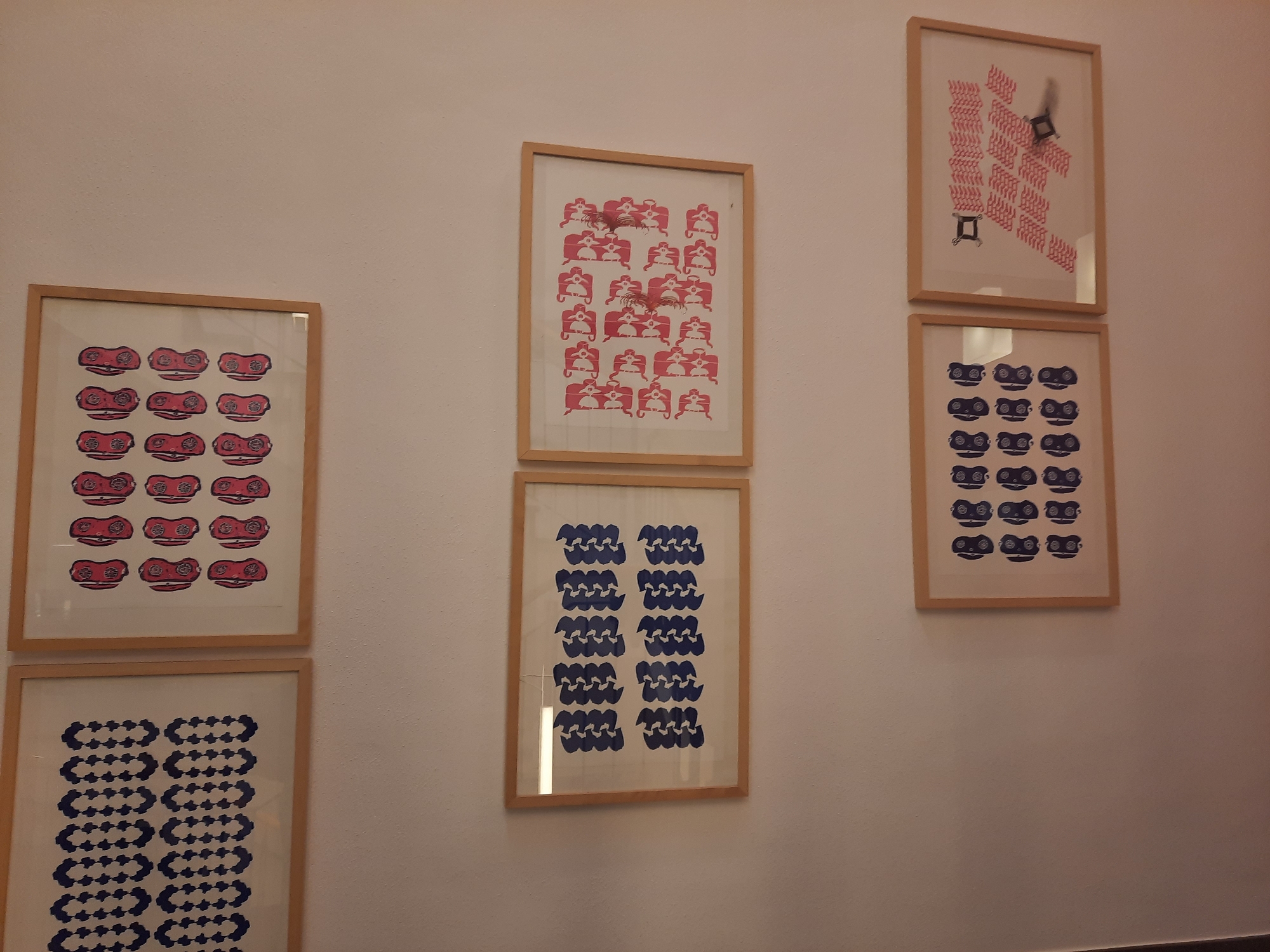 A medium shot shows a wall with six framed artworks. The artworks are arranged in two columns and three rows. The two artworks on the left are vertically oriented and appear to be silk screen prints. The top left artwork is a grid of red objects with circular indentations. The bottom left artwork is a grid of blue abstract shapes arranged in rows. The two artworks in the center are also vertically oriented and appear to be silk screen prints. The top center artwork features many small red figures sitting on chairs. The bottom center artwork is a grid of blue wavy shapes. The two artworks on the right are also vertically oriented and framed. The top right artwork is a grid of red diagonal lines with two small black objects. The bottom right artwork is a grid of blue objects that resemble goggles. The overall lighting suggests an indoor setting, possibly an art gallery or a private residence, with a plain white wall as the background. There are no people or animals visible in the image. The artworks are hung at varying heights and spaced evenly. The image was taken in Rosenheim, Germany.