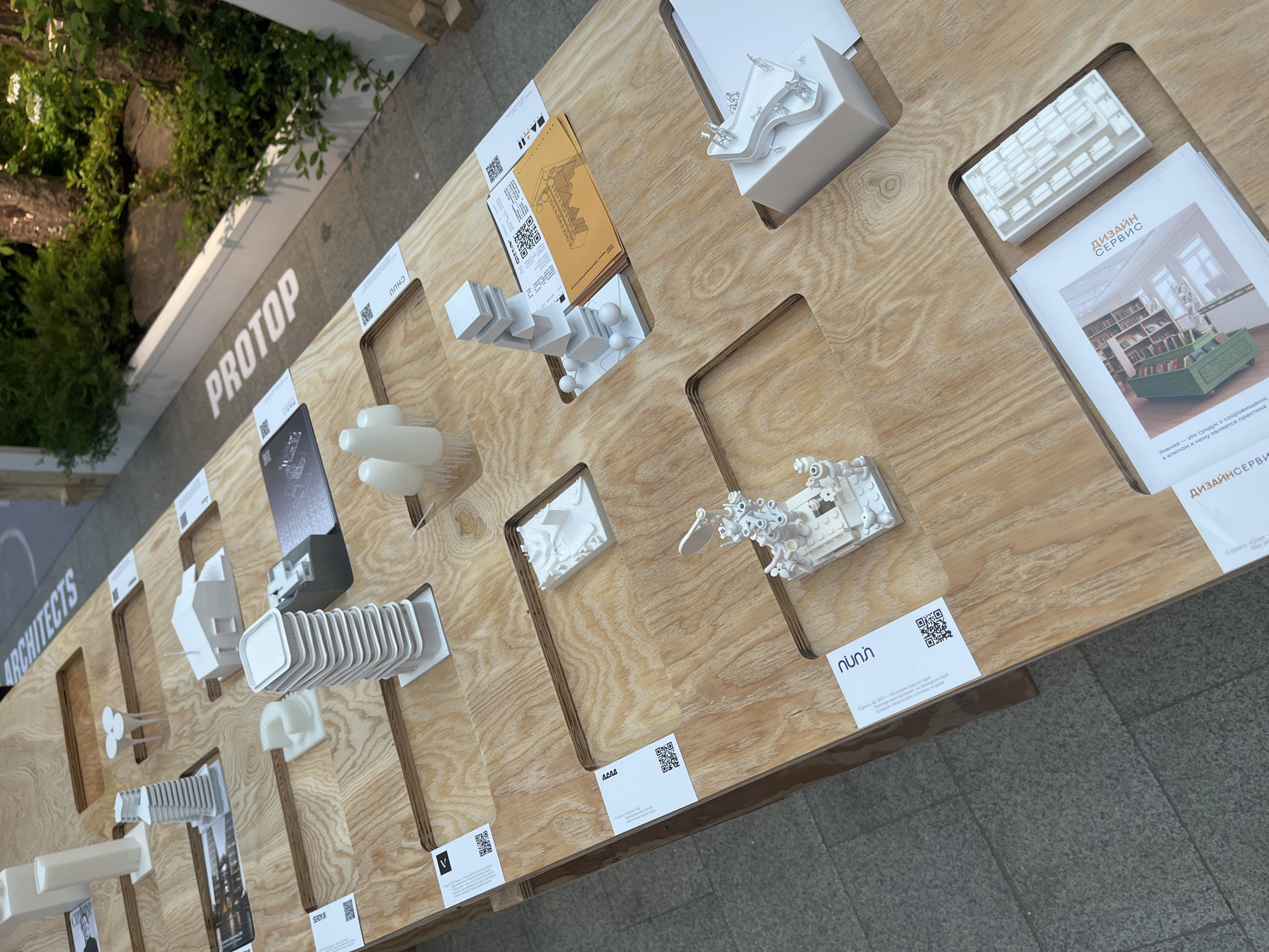 The image depicts a wooden display table with architectural models, printed materials, and signage, likely part of an exhibition, fair, or conference related to architecture and design. The table is positioned outdoors, as suggested by the view of trees and greenery in the background. The signage displays the word "PROTOP" followed by "ARCHITECTS," indicating the name of an organization or exhibition. The various architectural models displayed are small, white, and represent different designs, possibly prototypes or conceptual projects. Accompanying each model is a printed card with information about the design or the architect, including QR codes. A brochure with the title "ДИЗАЙН СЕРВИС" (Design Service) is also present. The overall scene suggests a presentation or showcase of architectural designs, possibly in Moscow, Russia, indicated by the Cyrillic text on the brochure and some signage.