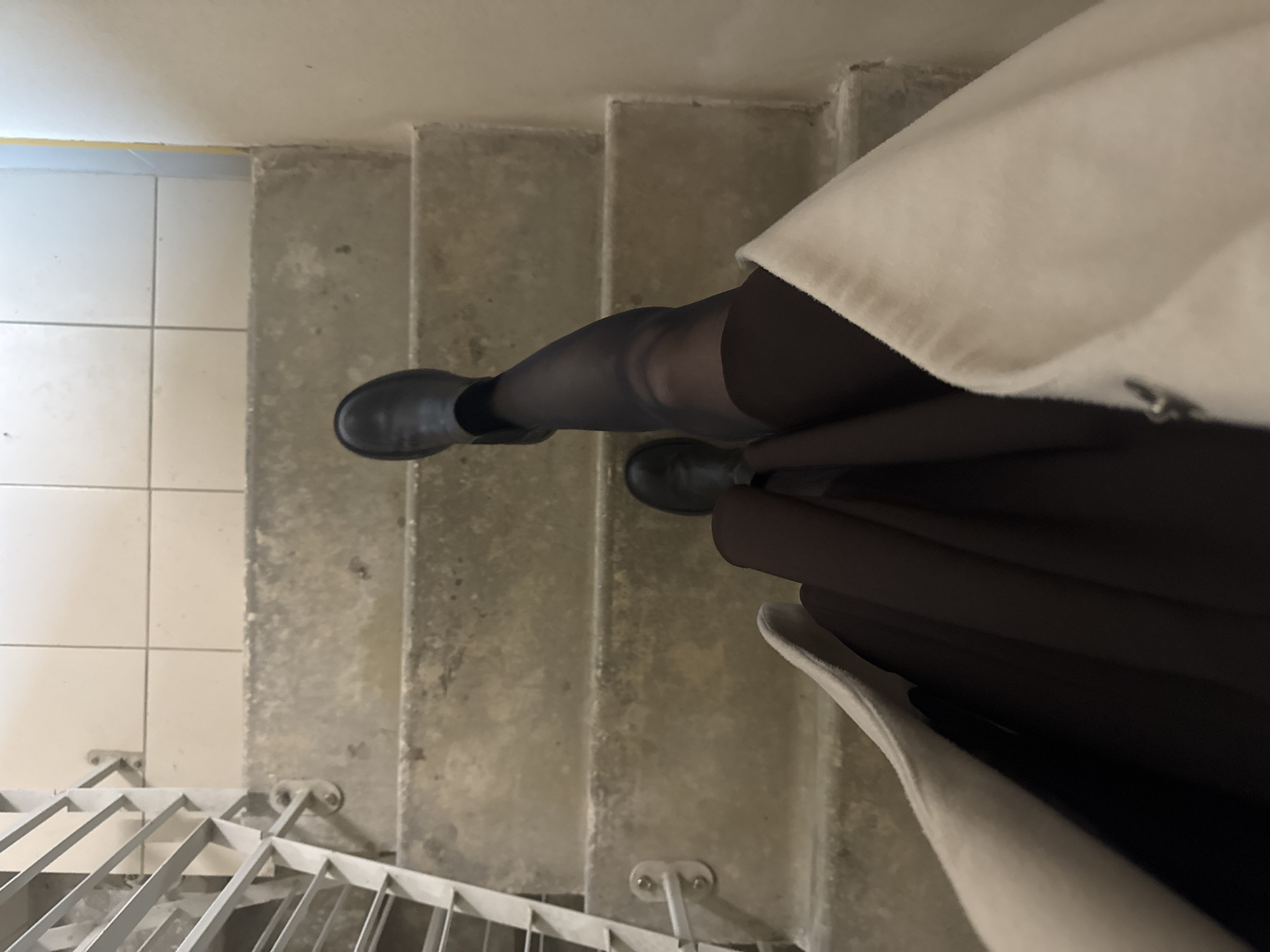 A top-down perspective image shows a person on a set of concrete stairs. The person wears black boots and black tights. A cream-colored coat is also visible. The stairs are likely located in an interior space, with a tiled floor visible on the left. A metal railing is at the bottom left. The specific location in Perm, Russia, cannot be determined from the visual information.
