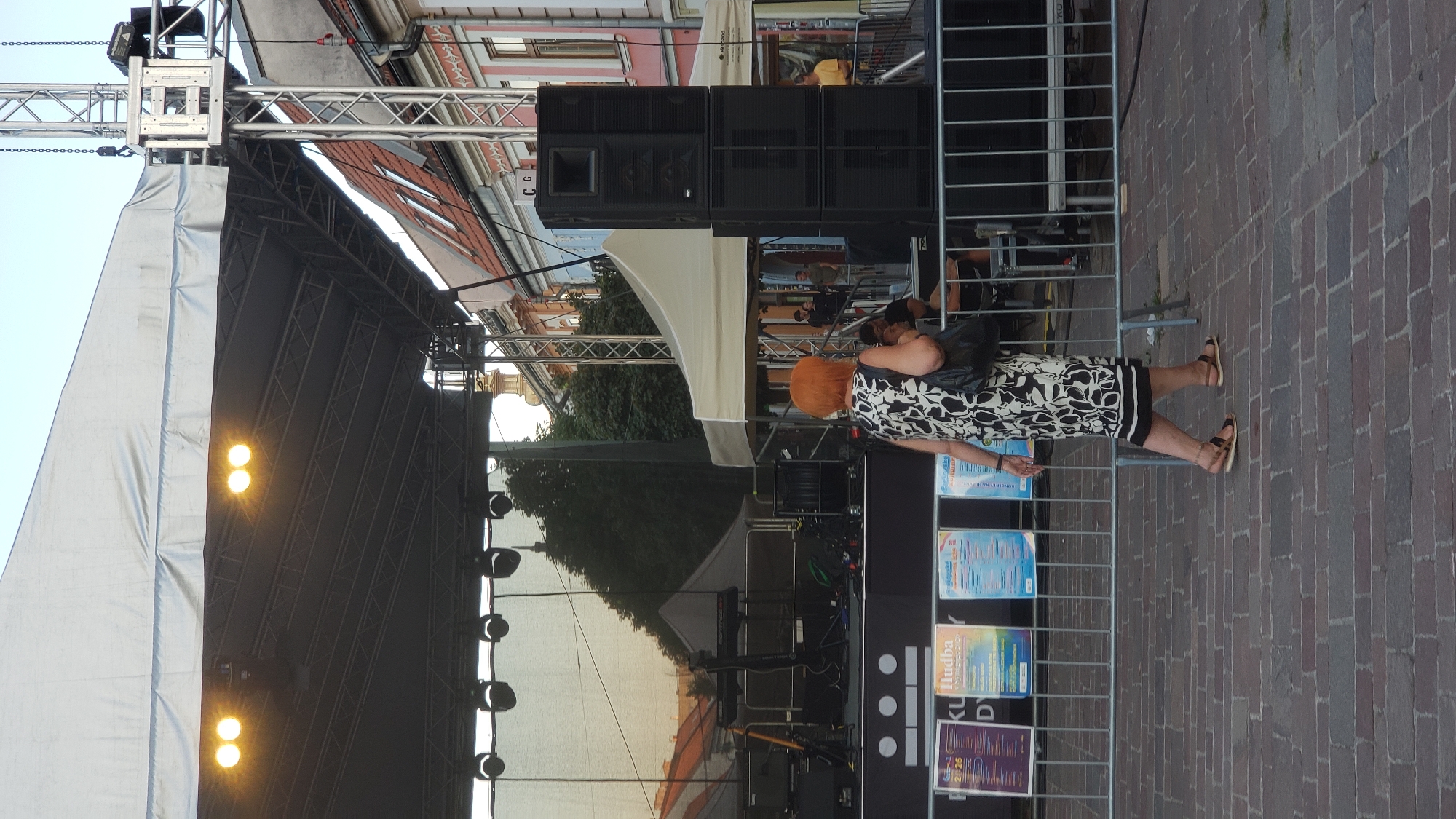 The image depicts an outdoor stage setup with a large white canopy. In the foreground, a woman with bright orange hair, wearing a black and white patterned dress and sandals, walks from right to left. She carries a black handbag over her left shoulder. Behind her, a metal barricade separates the public area from the stage. Several posters are visible attached to the barricade, including one with the text "Hudba" (music in Slovak). The stage itself has lighting equipment and audio speakers stacked vertically on the right side. In the background, buildings with red tiled roofs line a street, suggesting an urban setting. The overall atmosphere appears to be of an outdoor event or festival.
