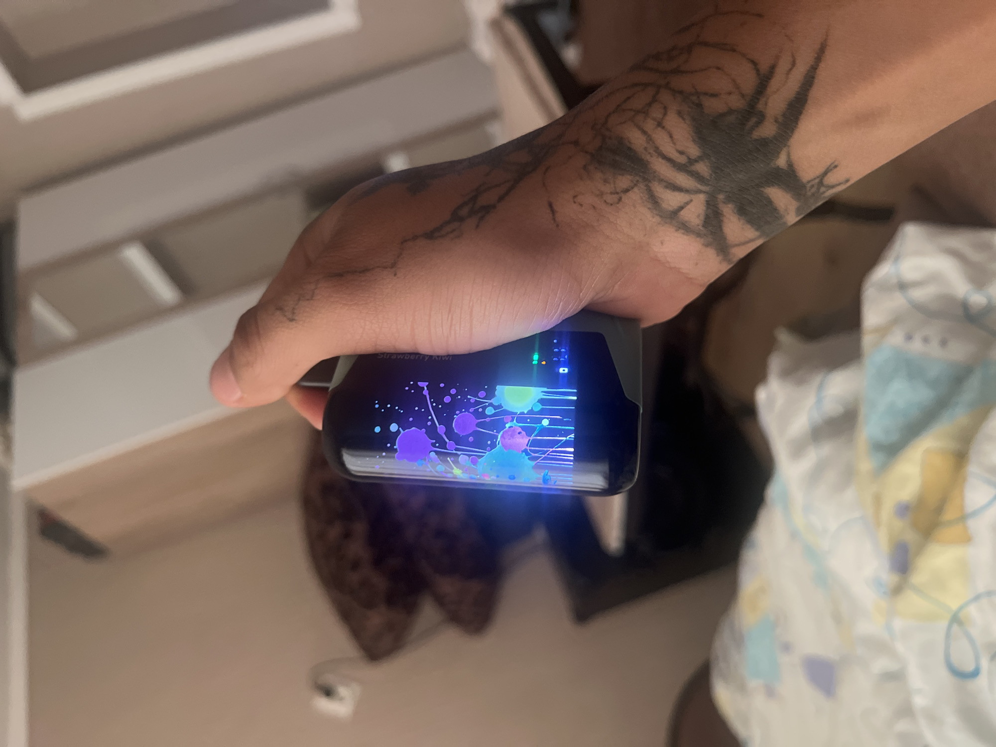 The image captures a close-up view of a person's right arm and hand holding a small, dark electronic device with a brightly illuminated screen. The person has a medium skin tone, and a large, dark tattoo covers much of their forearm, depicting an abstract, branch-like or splintered design. The device's screen displays a vibrant, abstract graphic featuring numerous colorful splatters and lines in shades of purple, blue, green, and pink, resembling paint splashes. Faintly visible text on the screen reads "Strawberry Kiwi."
The background is softly lit and out of focus, showing light-colored, possibly beige or off-white, walls or furniture surfaces, indicating an indoor setting. On the right side of the frame, a soft, patterned fabric, predominantly white with light blue, yellow, and purple accents (possibly from a blanket or clothing), is visible. The glowing screen suggests the photo was taken in dim lighting conditions, likely evening or night. The scene is located in Sofia, Bulgaria.