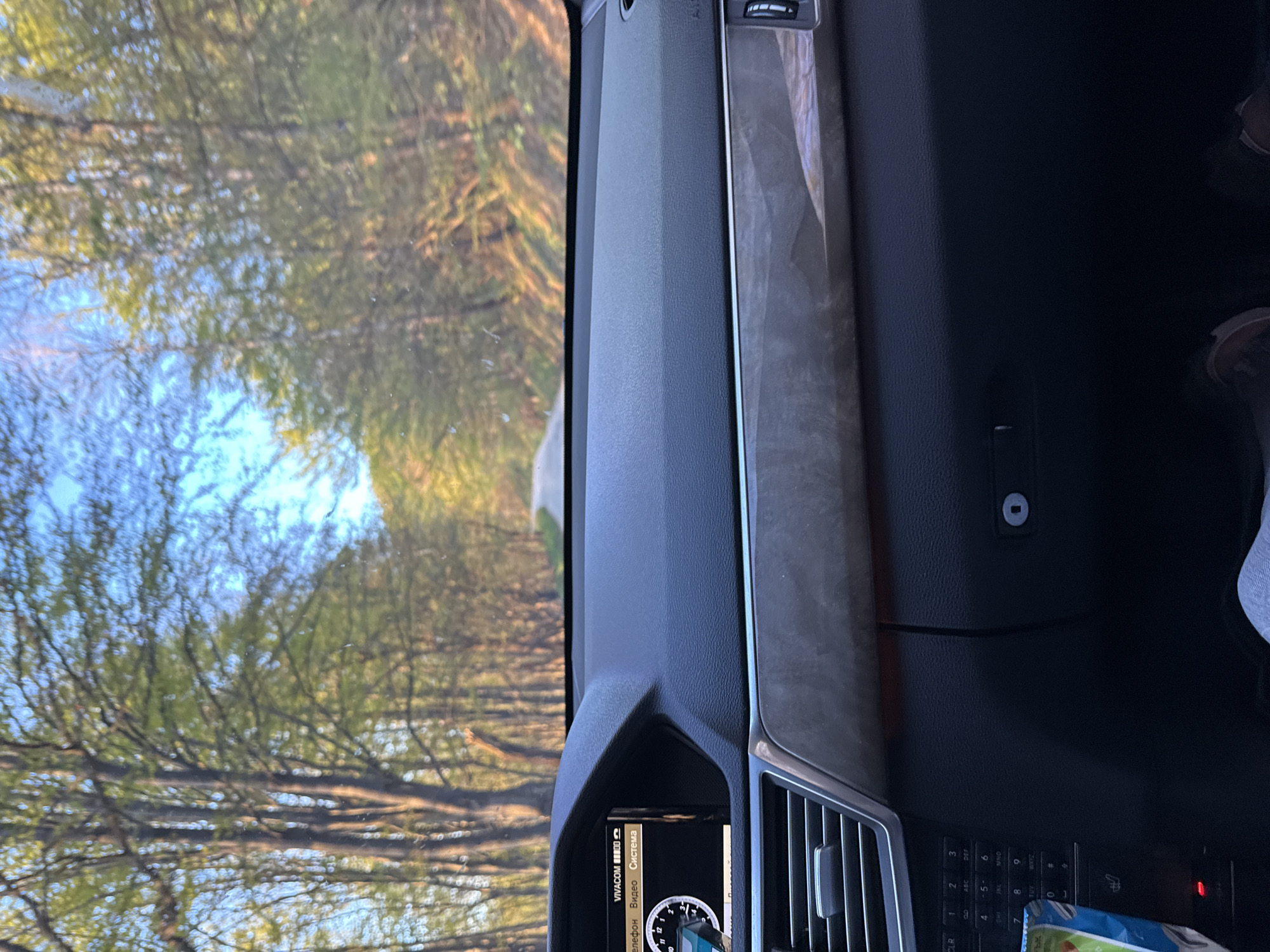 The image shows the interior of a car driving on a road lined with trees. The view is from inside the vehicle, looking out through the windshield. The car's dashboard and interior features are visible, including a navigation system or entertainment display, air vents, and some controls. There is the edge of what appears to be a passenger's leg. The reflection of the trees can be seen in the windshield.
The image is too indistinct to reliably pinpoint the exact location.