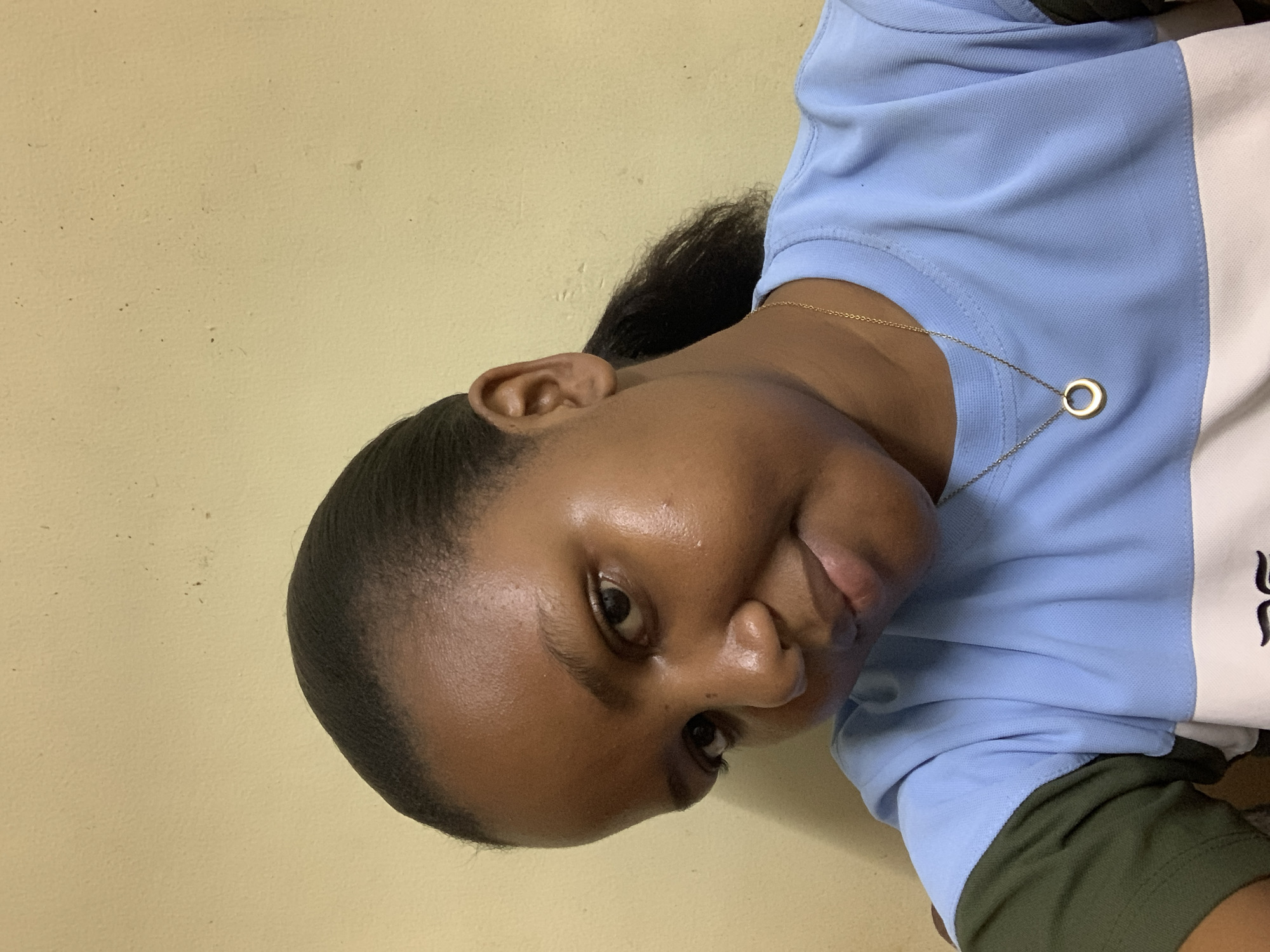This close-up, portrait-style photograph is rotated 90 degrees counter-clockwise, featuring a young person with dark skin. The subject, who appears to be female, looks directly into the camera with a calm expression and a subtle smirk. Her dark hair is pulled back into a ponytail, with some curly strands visible behind her right ear. Her skin exhibits a natural sheen, and her eyes are wide open.
She is wearing a light blue polo shirt with a distinct white vertical stripe down the right side of the torso (from the viewer's perspective). A piece of dark olive green fabric, likely a sleeve or an undershirt, is visible underneath the blue. Around her neck, she wears a thin gold chain necklace with a small, circular gold pendant.
The background is a plain, light beige or pale yellow wall, which shows minor specks and texture, suggesting an indoor setting. No specific location cues, time of day, weather, or visible text are present in the image. The overall mood conveyed is casual and direct.