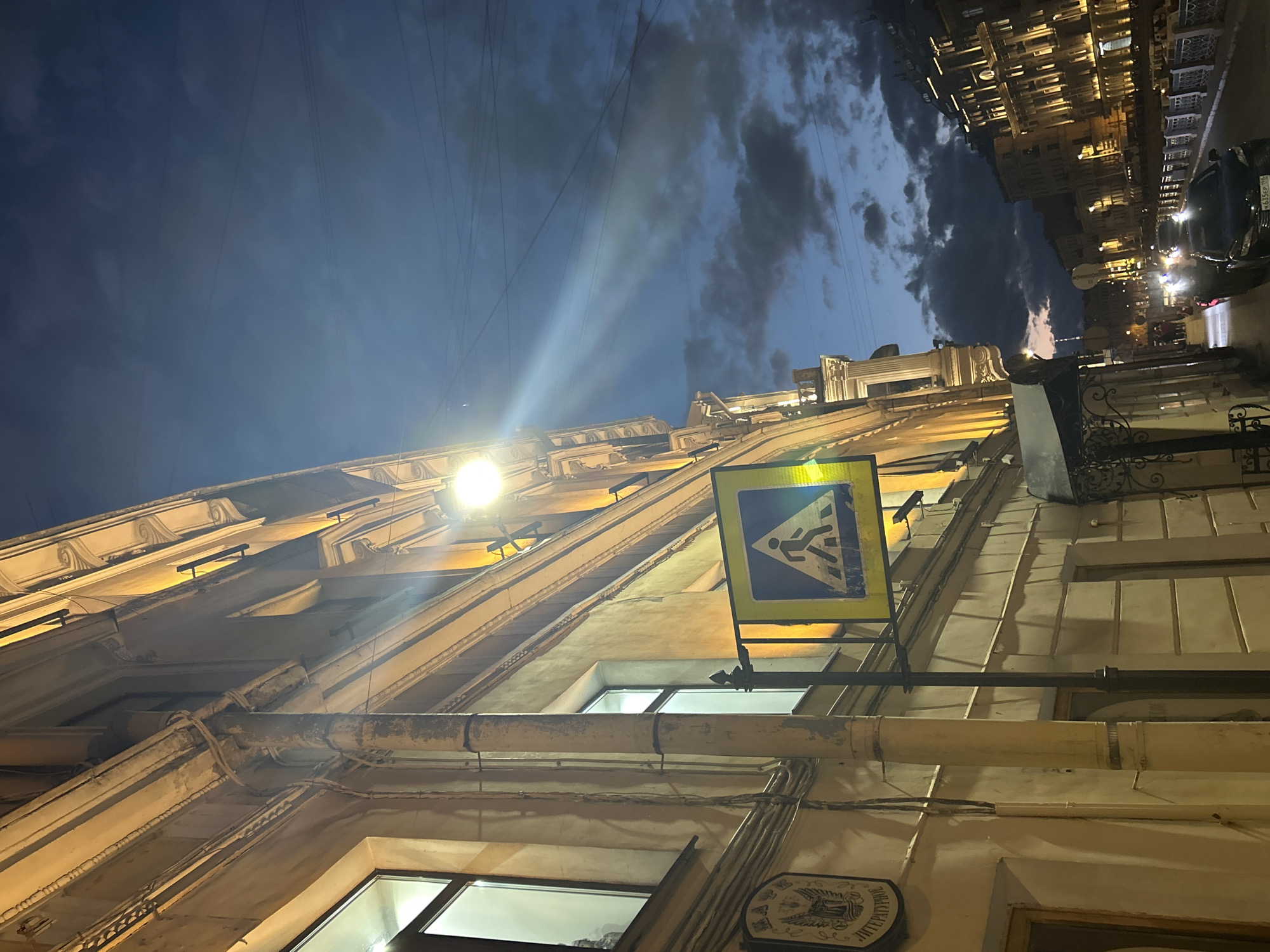 The image captures a street scene at dusk in an urban environment. On the left, a building with classical architectural details is illuminated by warm yellow lights. A downspout runs vertically along the building's facade. A yellow and blue pedestrian crossing sign is prominently displayed, mounted on a dark pole. Above, the sky is a deep blue, transitioning from twilight to night, with scattered clouds and overhead wires visible. To the right, another building with illuminated windows lines the street, and further down, more buildings with lit facades and decorative elements can be seen. Cars are parked and driving on the street, and there are a few indistinct figures walking on the sidewalk. The overall impression is a moody, atmospheric urban evening.
