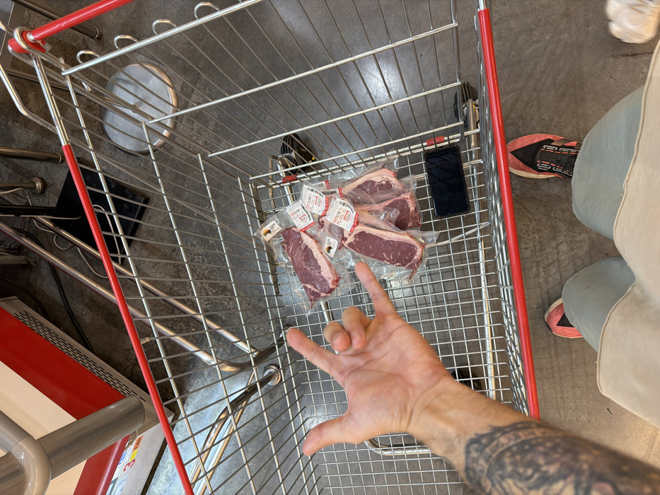 The image provides a top-down view into a metal shopping cart, situated within a supermarket in Bangkok, Thailand. The cart contains multiple vacuum-sealed packages of raw red meat, identified as steaks or chops, each bearing a white label with red text indicating pricing in Thai Baht. A black smartphone is visible lying flat on the cart's floor. A person's left arm, visibly tattooed, extends from the lower left of the frame, with the hand performing a "rock on" gesture (index and pinky fingers extended). The lower portion of the individual's body is visible, clad in light-colored trousers or shorts and dark athletic shoes with red accents. The surrounding environment features an industrial-style concrete floor, with a portion of a white and red commercial refrigeration or display unit visible in the upper left background, along with other miscellaneous metal fixtures.