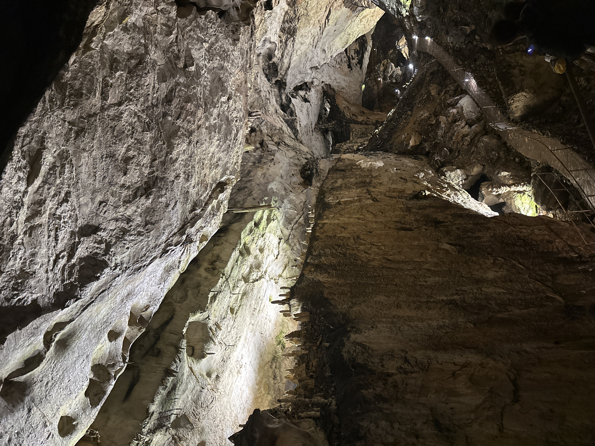 The image depicts the interior of Magura Cave, located near Raianovtsi, Bulgaria. The view is upward, showcasing the cave's walls and ceiling. The cave walls are primarily composed of light-colored rock formations with visible geological features like ridges and depressions. A constructed walkway is visible near the top of the image, suggesting human modification for tourism or exploration. The lighting is a combination of natural and artificial, casting shadows and highlighting the texture of the rock surfaces. The color scheme is dominated by shades of gray, white, and brown.
