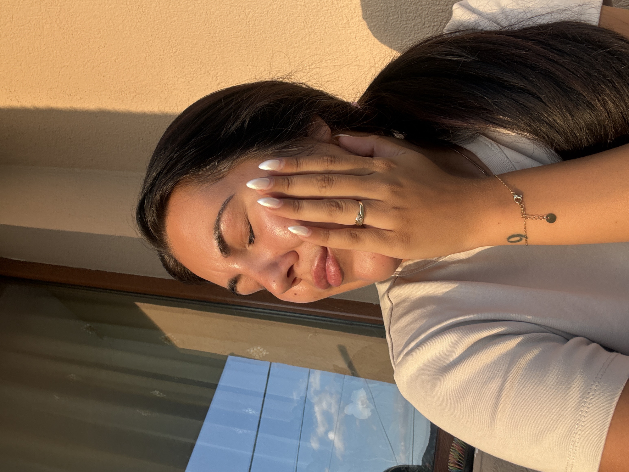 A close-up, medium shot captures a young woman from the chest up, positioned in front of a beige wall and a window. The woman, with her eyes closed and hand gently touching her cheek, is bathed in the warm glow of what appears to be sunset. Her dark hair is pulled back into a ponytail. She wears a light-colored t-shirt, and her fingernails are painted white. A gold ring with a small stone is visible on her left hand, as is a delicate gold bracelet with a circular charm and a tattoo of the number "9" on her right forearm. The window reflects the blue sky with clouds, suggesting an outdoor setting. The image was taken in Strelcha, Bulgaria.