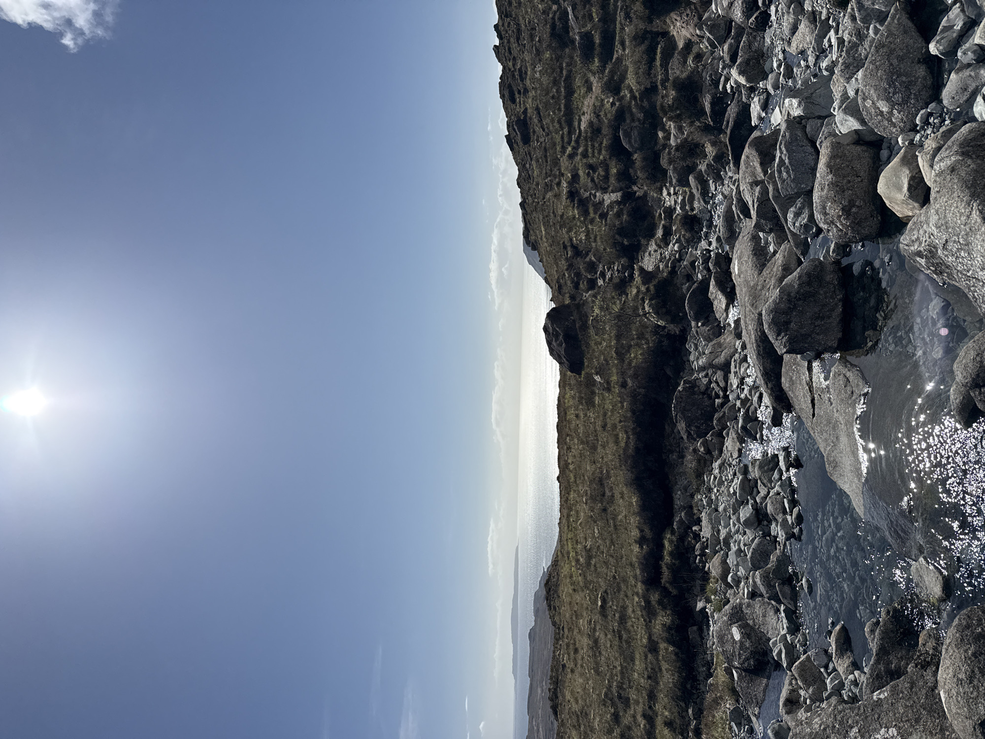A landscape photograph shows a sunlit sky transitioning to a bright horizon over the sea, adjacent to a rocky terrain covered with grass and a small stream. The sun is positioned in the upper left corner, casting a strong glare. The rocky terrain is densely populated with stones of varying sizes, some of which are partially submerged in the water. The environment suggests a natural setting, potentially a coastal area near a hill or mountain. There are no people visible in the image. The location is specified as Unknown, Unknown.