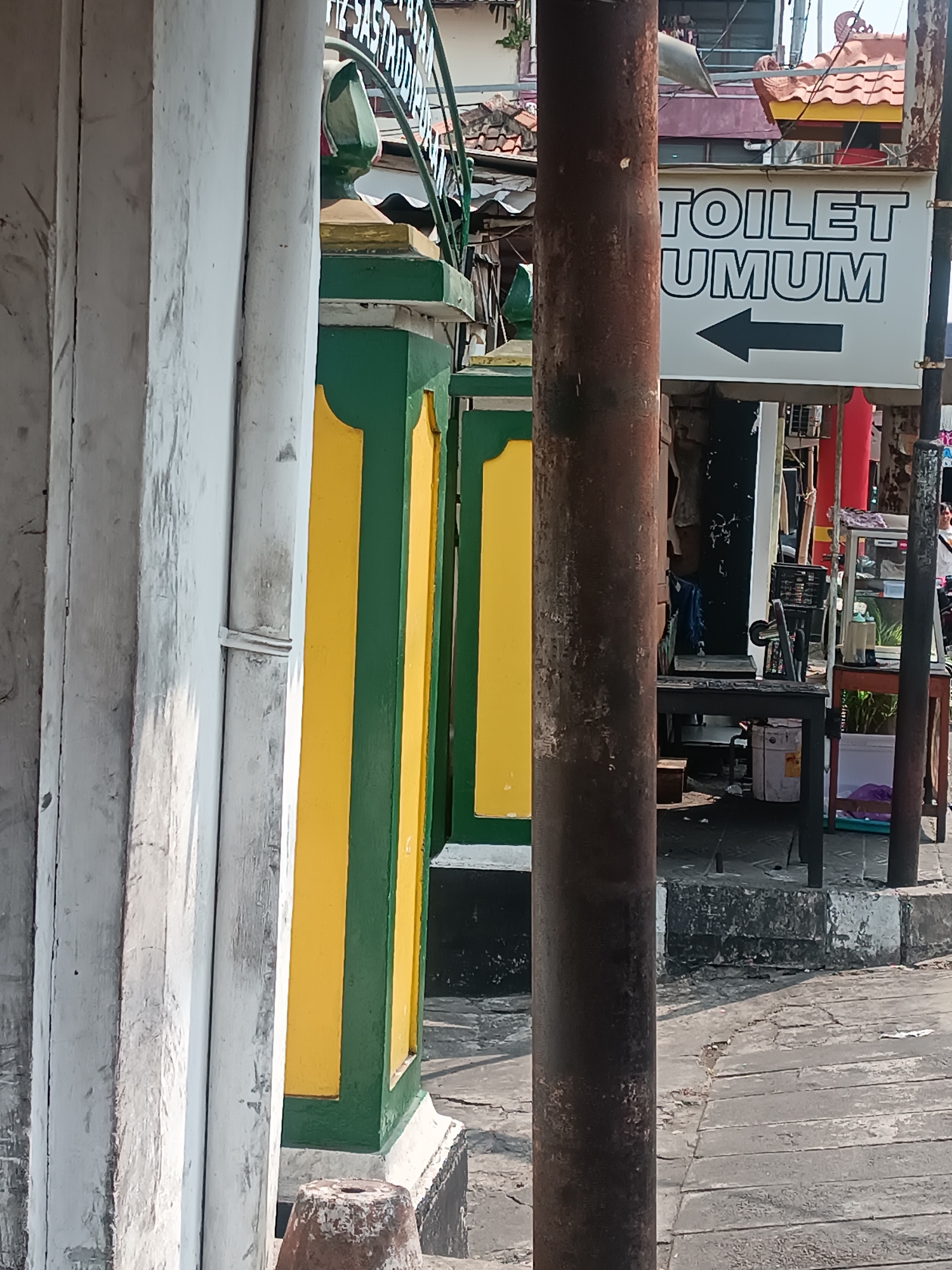 A street scene in Yogyakarta, Indonesia, features a signpost with the words "TOILET UMUM" and an arrow pointing left, indicating a public restroom. The sign is white with black lettering and a black arrow. In the foreground, a rusty metal pole stands vertically, obscuring part of the scene. To the left of the pole, a section of a building with white painted wooden pillars is visible. A green and yellow striped column also stands to the left, suggesting an architectural element of a building or structure. In the background, several stalls or shops are present, with some merchandise displayed, including what appears to be a sewing machine on a table. The ground is paved with cracked concrete slabs. The overall environment suggests an outdoor commercial or public space.