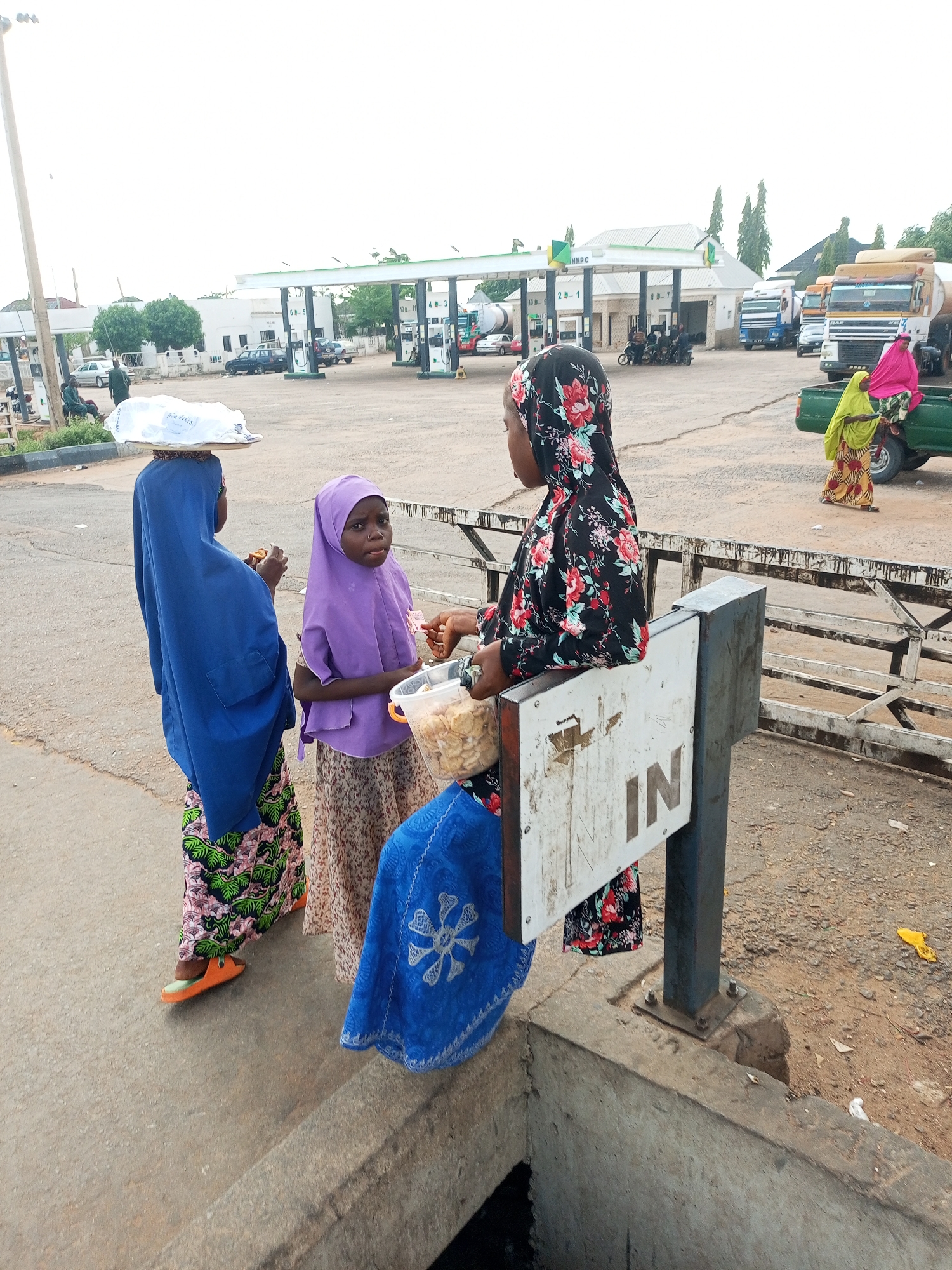 The image captures an outdoor scene at what appears to be a fuel station or depot. In the foreground, three young females are interacting. One, on the left, wears a bright blue hijab and a patterned skirt, carrying a tray of items balanced on her head. Beside her, a girl in a purple hijab is holding a plastic container filled with snacks. To the right, another girl, dressed in a black hijab with a floral pattern, leans against a metal post supporting a sign that reads "IN." She is also holding a container with snacks. In the background, fuel pumps and station buildings are visible, along with several large trucks and other vehicles. The ground is a mixture of pavement and dirt. The sky is overcast.