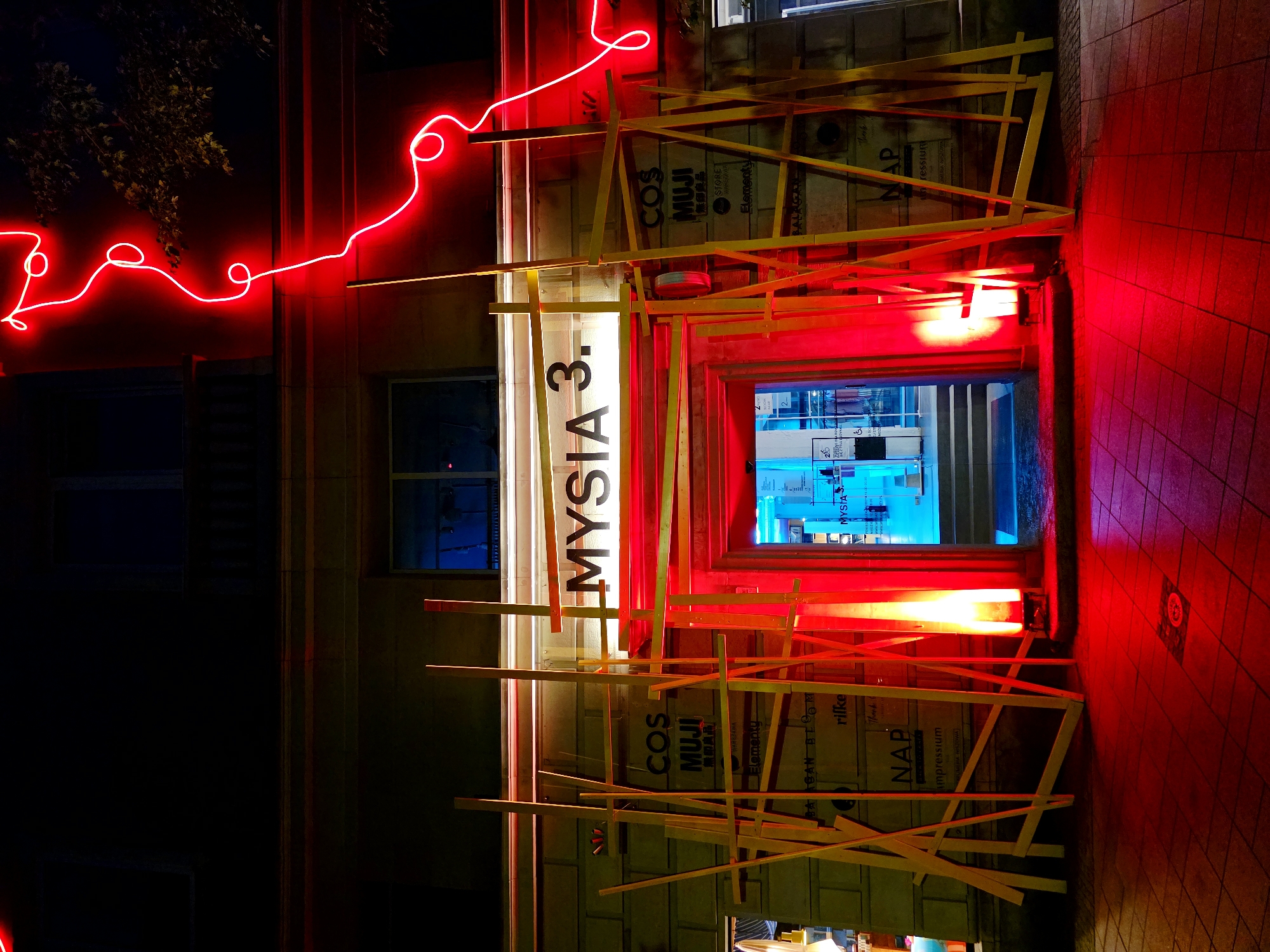 This is a nighttime, outdoor, vertical photograph of the exterior of a building in Warsaw, Poland, illuminated by red neon lights and a bright blue light emanating from an interior doorway. The scene is abstract and somewhat chaotic, with a haphazard construction of light-colored wooden beams crisscrossing in front of the building's facade. These beams obscure parts of the building's signage and entrance.
The building itself appears to be a commercial establishment. Visible text on the facade and within the doorway includes brand names such as "COS," "MUJI," and "MYSIA 3." Other text snippets like "WARSZAWA," "Elementy," "BAAGAN BLOOM," "NAP," and "rifke" are also present, suggesting a retail or gallery space. The red neon light snakes across the upper part of the building, adding a vibrant, almost electric glow to the dark surroundings. The ground in front of the building is a red-tiled walkway, reflecting the red light and creating an intense, saturated atmosphere. The overall impression is one of an art installation or a temporary structure, with a stark contrast between the organic, artistic arrangement of the wooden beams and the modern, commercial branding. The time of day is clearly night due to the artificial lighting and dark background, and the weather appears to be clear, as evidenced by the sharp reflections.