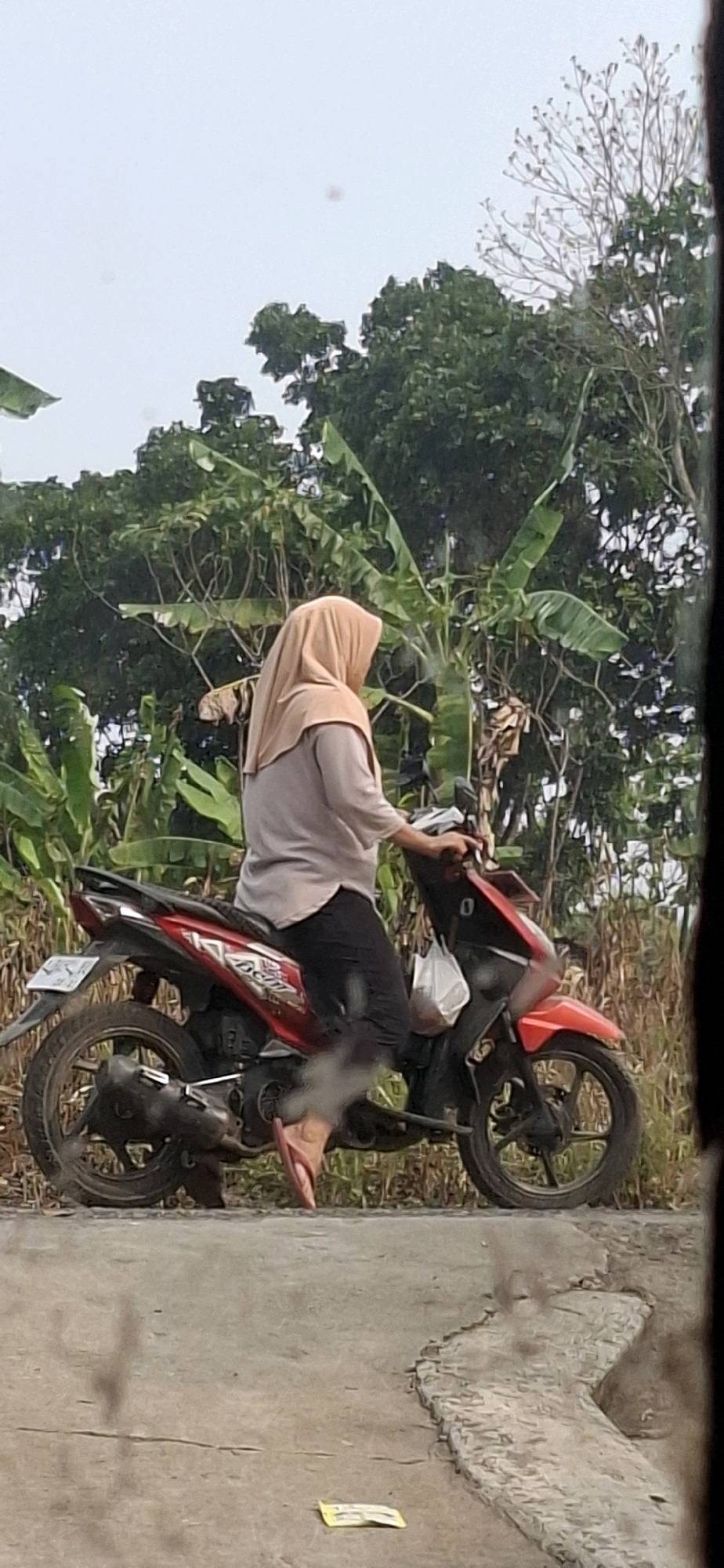 A woman wearing a beige hijab and a light grey shirt is seated on a red and black motorcycle, facing to the right. She is wearing black pants and pink flip-flops. The motorcycle is parked on the side of a paved road, with a dirt shoulder and vegetation behind it. The vegetation appears to be tropical, with large leaves and trees. The sky is overcast. The scene is in Cibinong Hilir, Indonesia.