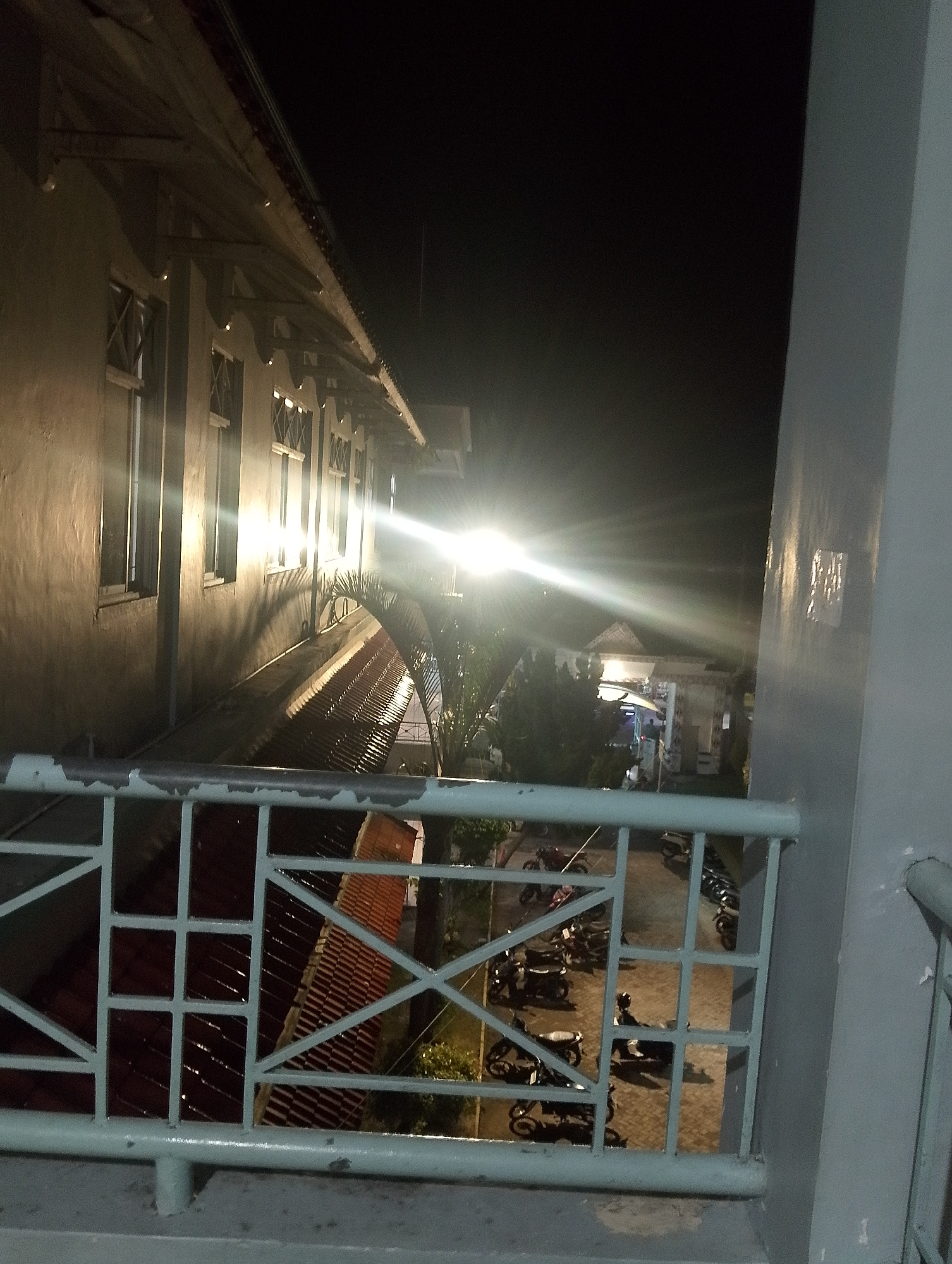 This image captures a nighttime view from a balcony overlooking a street in the City of Pematang Siantar, Indonesia. A light source, possibly a streetlamp, creates a strong glare and lens flare in the center of the frame, illuminating the side of a building on the left and casting shadows. The building has several windows with a lattice design. Below the balcony, a roof with red tiles is visible, and beyond that, a street lined with parked motorcycles. The scene is dark, indicating it is nighttime, and the lighting suggests an urban or semi-urban environment.