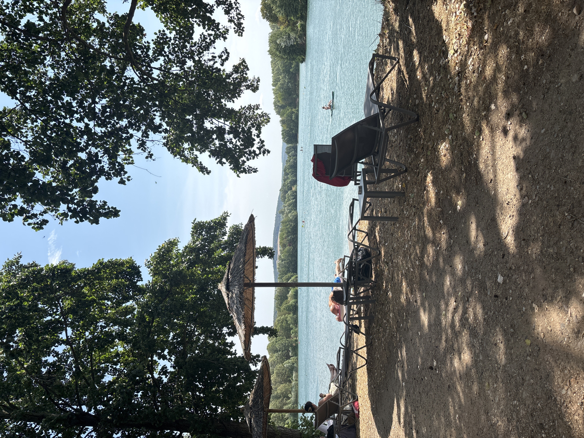 This image depicts a sunny day at a lake in Niedernberg, Germany. In the foreground, a sandy shore is lined with empty lounge chairs and two thatched umbrellas, offering shade. The lake itself is calm and turquoise, with several people visible enjoying various water activities. In the distance, a treelined shore forms the horizon. The overall scene conveys a peaceful, recreational atmosphere.