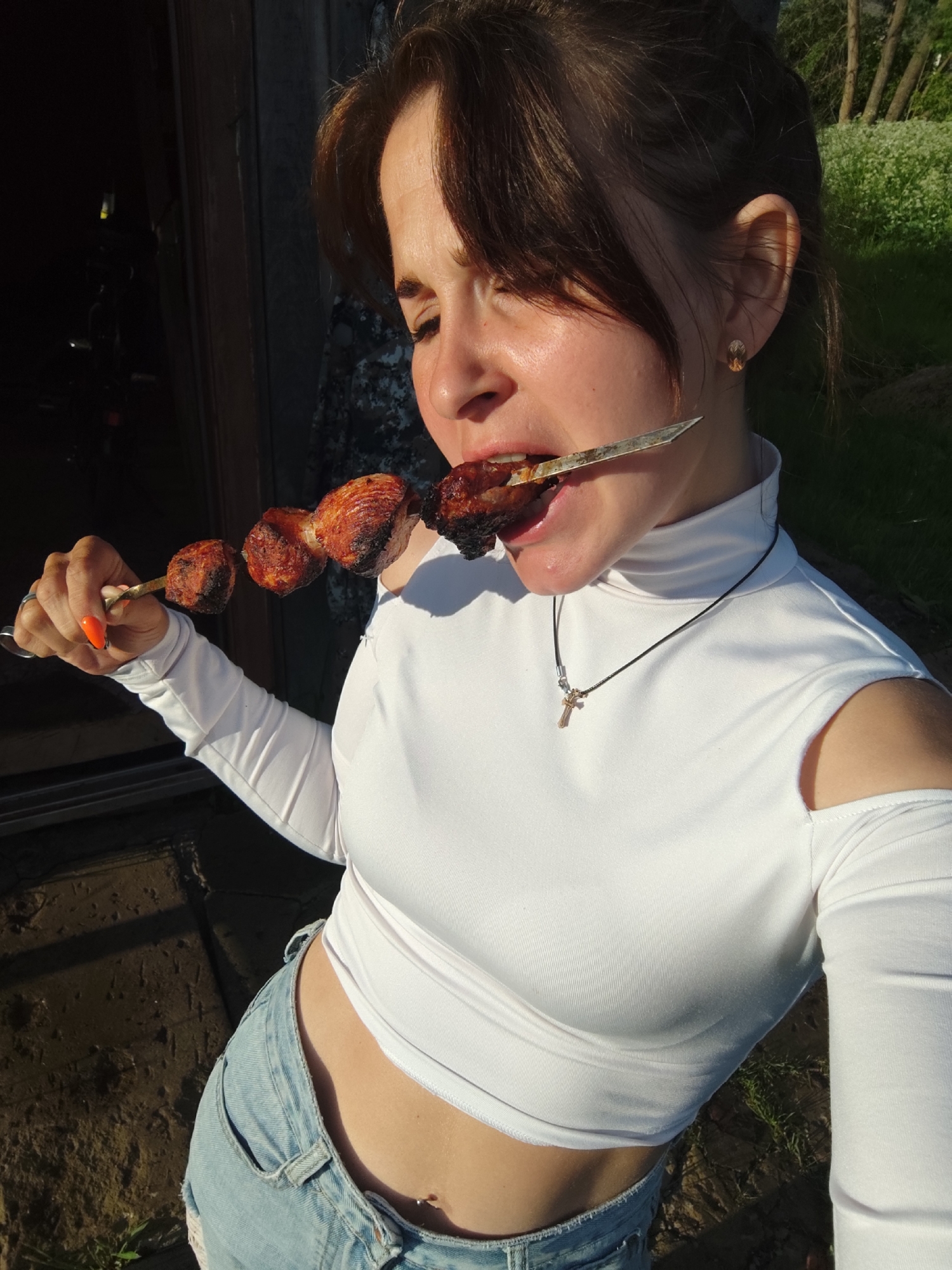 A young woman is captured outdoors in Фоки, Russia, on a bright, sunny day, engaged in eating grilled meat from a skewer. She has dark brown hair, with some strands framing her face, and her eyes are partially closed, possibly squinting from the sun or in enjoyment. She wears a white, form-fitting long-sleeved crop top with an off-the-shoulder design and a high neck, paired with light blue denim shorts or jeans. A dark cord necklace with a small gold-colored cross pendant adorns her neck. She also has a small gold-colored earring in her right ear and a tiny belly button piercing. Her fingernails are painted a vibrant orange.
She holds a metal skewer laden with several pieces of darkly charred and caramelized grilled meat, which she is biting into. The meat appears to be well-cooked, resembling shashlik or kebab. The strong sunlight casts distinct highlights on her skin and clothing, and there are visible shadows.
The background on the left features a dark, rustic structure, possibly a wooden building or door frame, suggesting an outdoor or rural setting. To the right, lush green grass and trees are visible under the bright sunlight. No visible text is present in the image.
