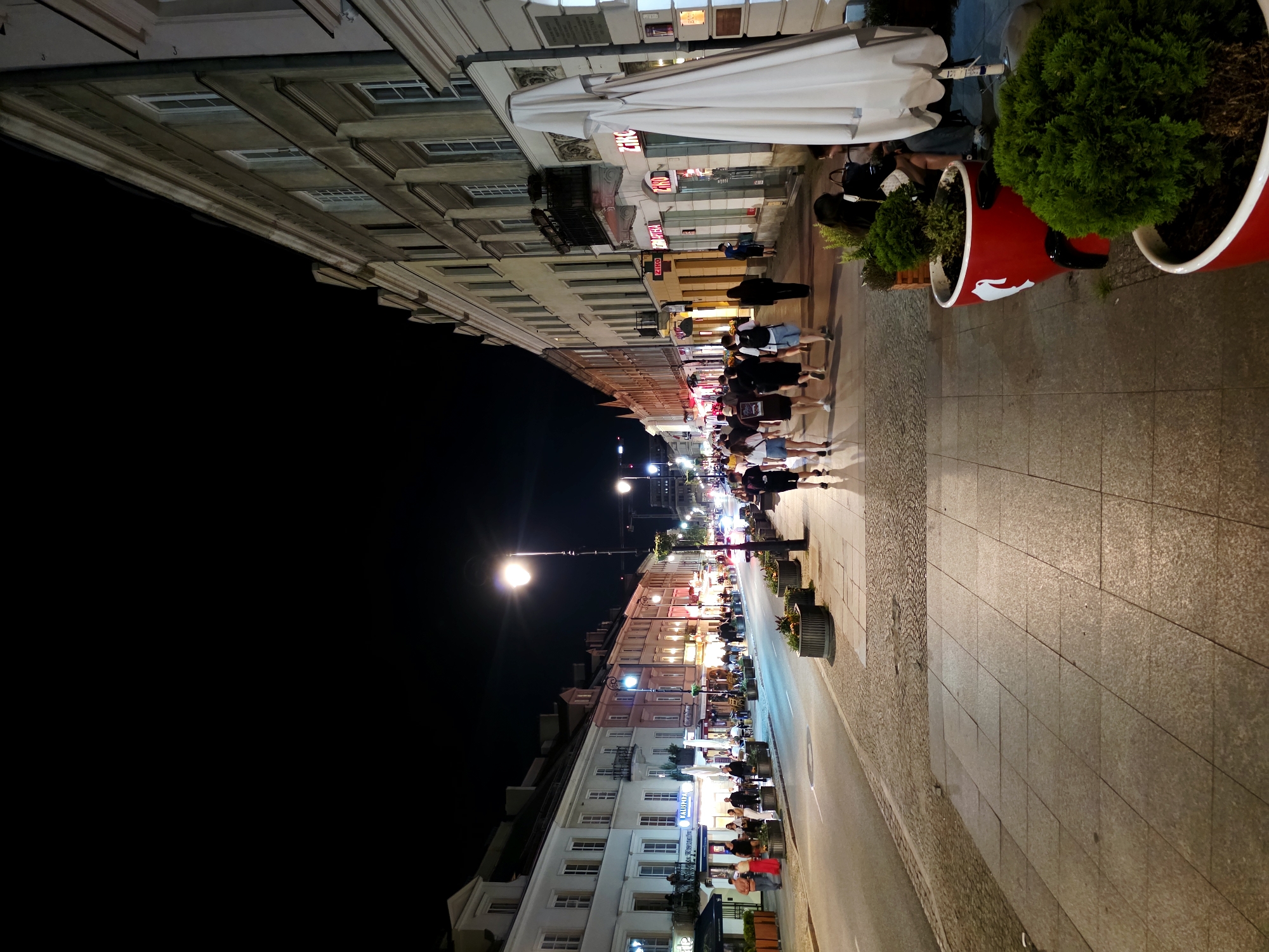 The image depicts a bustling street scene at night, likely in Warsaw, Poland, given the provided context. A wide cobblestone and tiled sidewalk runs alongside buildings that house shops and restaurants. Several people are visible, walking along the sidewalk and seated at outdoor tables, suggesting a lively evening atmosphere. Some individuals are gathered in groups, while others walk alone.
The buildings feature traditional architecture with multiple windows and ornate detailing. The street is illuminated by streetlights and the warm glow emanating from the shops and restaurants, creating a welcoming ambiance. Large planters containing green shrubbery are placed along the sidewalk, adding a touch of nature to the urban setting. One planter, a vibrant red cylindrical container with a white logo, stands out.
A large white awning extends from one of the buildings, providing shade or shelter. Visible text includes what appears to be shop signage with lettering such as "ZNAK" and "TAU..." on the building facades, illuminated by neon lights. The time of day is clearly nighttime, and the weather appears to be pleasant enough for outdoor dining and strolling. The overall mood is one of activity and enjoyment, typical of an urban evening.