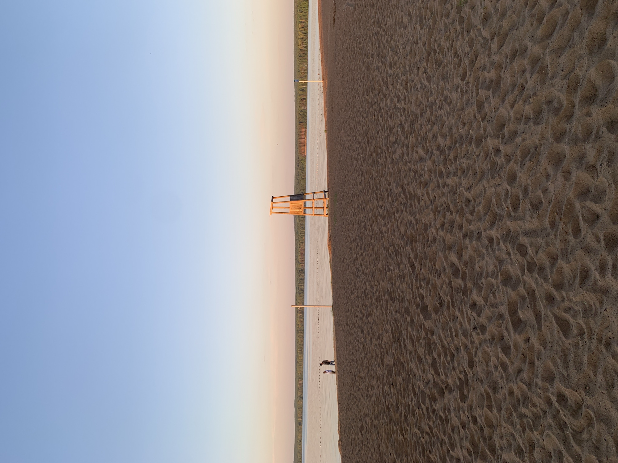 The image depicts a beach at sunset in Chaikovsky, Russia. The scene is divided into three main sections: a clear blue sky on the left, a sandy beach on the right, and a body of water in the middle. A lifeguard stand is positioned along the waterline. Two people are seen walking along the shoreline near the center of the frame. The sand exhibits textural details with numerous small depressions and ripples. The background shows a tree line.