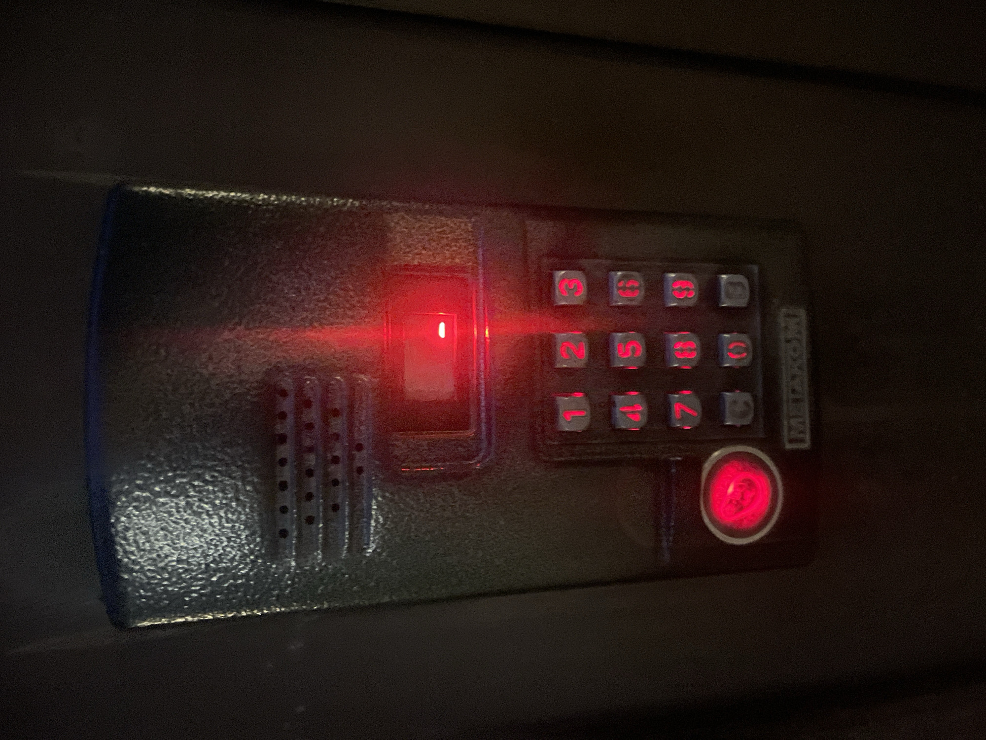 A close-up shot shows a weathered, dark gray intercom panel mounted on a dark background. The panel has a textured surface and features a speaker grille at the top. Below the grille is a rectangular slot with a red light emanating from it. Beneath this are illuminated red numeric buttons arranged in a standard keypad layout, from 1 to 9, with 0 and a star or pound key at the bottom. To the left of the numeric keypad is a circular red button, which is also illuminated. The word "METAKOM" is visible in a silver plate at the bottom of the panel. The overall impression is that of a functional, if somewhat aged, entry system. The location is identified as Markovsky, Russia.