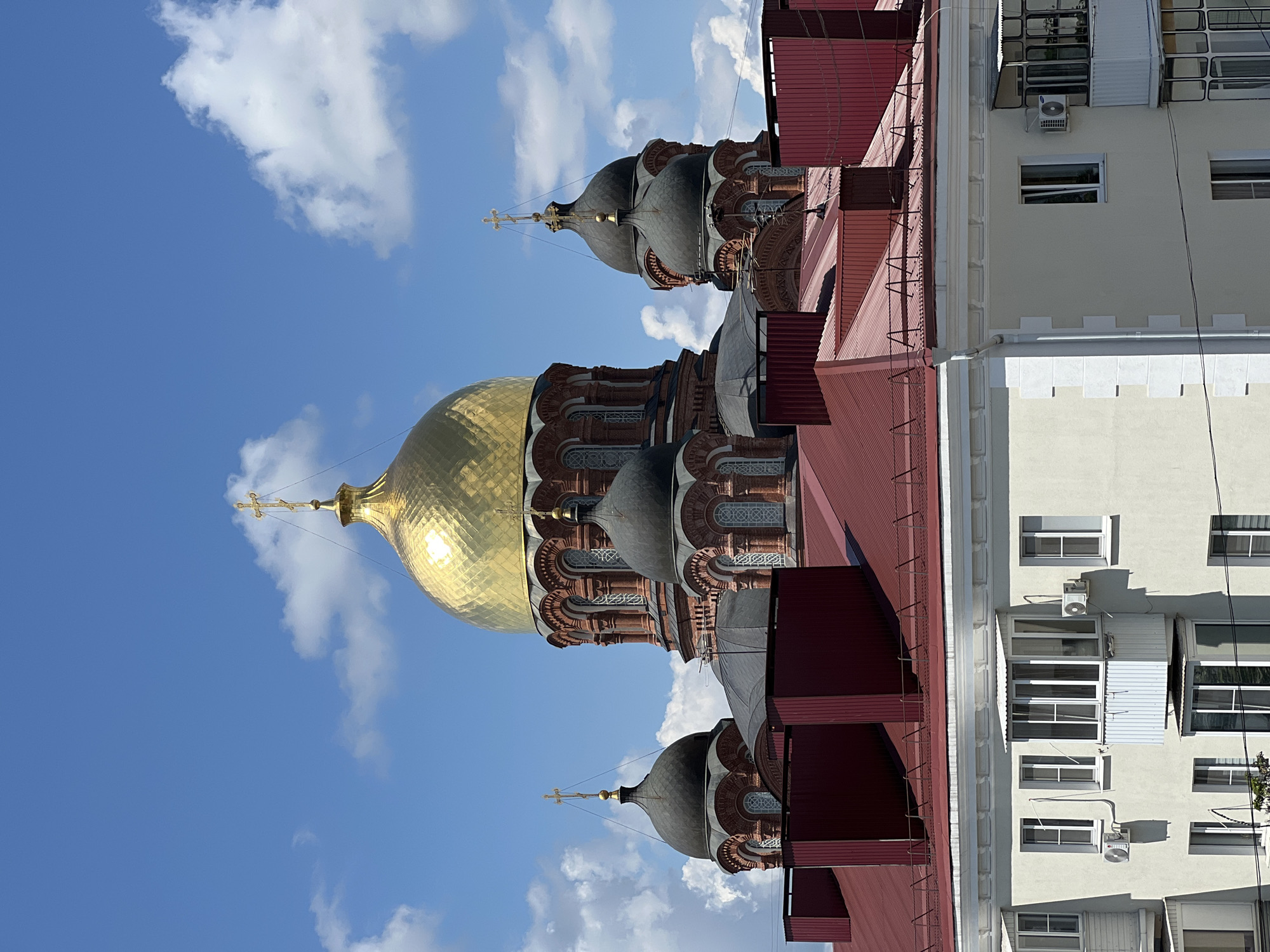 The image shows an aerial view of the Cathedral of St. Catherine in Krasnodar, Russia. The grand cathedral, constructed of red brick, is adorned with multiple domes. The central dome is a prominent golden color, while other domes are dark gray. The cathedral is surrounded by a series of smaller domes, also dark gray, each topped with a cross. In the foreground, a modern apartment building with a red metal roof is visible. The building has several windows and balconies. The sky is blue with scattered white clouds.