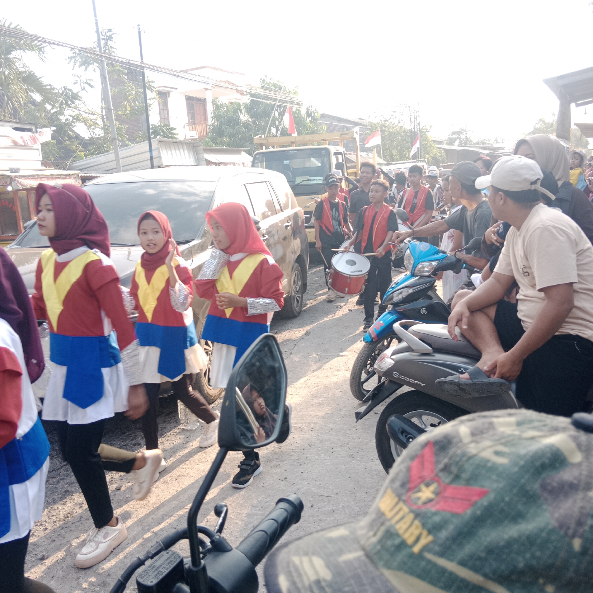 The image captures a street scene in Tegalrejo, Indonesia, likely during a daytime event or parade. In the foreground, a person wearing a camouflage hat with a "MILITARY" patch is partially visible, looking to the right. A motorcycle with a black handlebar is also in the immediate foreground.
The main subjects of the image are a group of young people, appearing to be participating in a performance. Several girls in matching red, yellow, and blue uniforms, complete with hijabs, are walking in a procession. Two of them are holding silver-colored accessories on their wrists. Behind them, a group of boys, some in similar red and black vests, are visible. One boy is playing a drum.
The background reveals a street with several parked vehicles, including a dark-colored SUV or minivan on the left and a light-colored truck further back. A building with white walls and a tiled roof is visible in the distance, along with trees and utility poles. Indonesian flags are mounted on the truck, suggesting a national celebration or event. The ground is unpaved, a dusty road surface. The lighting indicates it is a bright, sunny day.