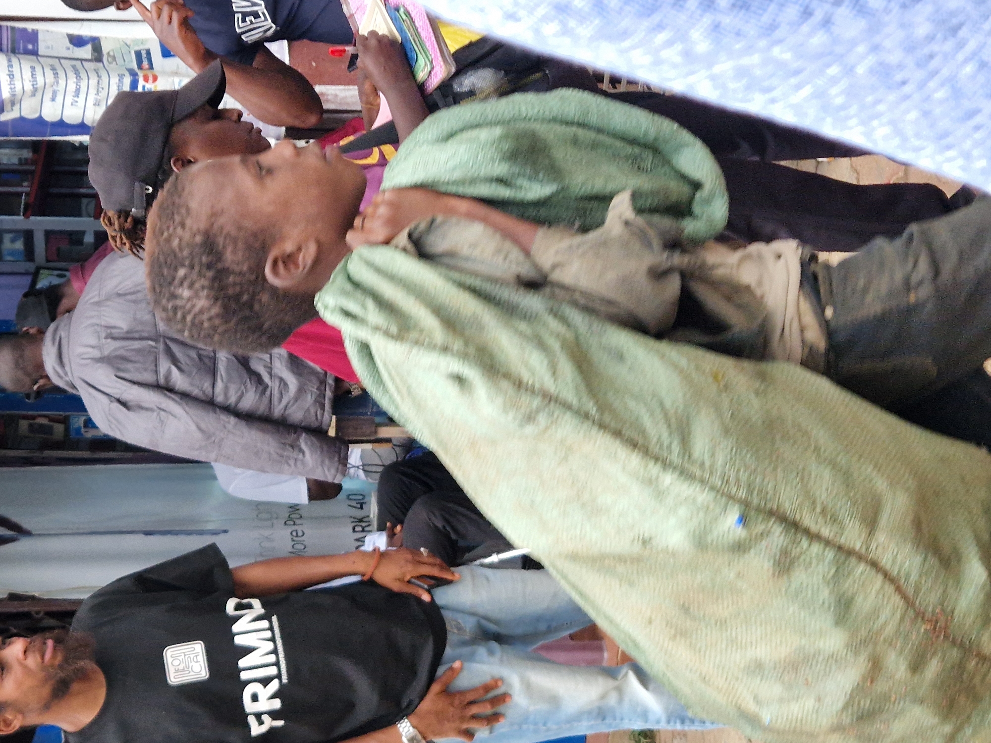 This media file depicts an outdoor scene in Jos, Nigeria, where several individuals are gathered. The foreground features a person wearing a black t-shirt with the white text "FRIMND" and a logo, and a red wristband. They appear to be looking upwards and to the side, with a slight smile. To their left, another individual is partially visible, wearing a grey, quilted jacket.
Further back, a person wearing a black baseball cap is seen from the side, facing away from the camera. A young person is wrapped in a light green cloth or blanket, also facing away. The background reveals a stall or shop with various items displayed, including what appear to be books or pamphlets, and possibly electronics or other goods. The overall setting suggests a bustling marketplace or street scene.
Visible text includes "FRIMND" on the t-shirt, and other partial text elements like "DUC," "Withdraw," "irtime," "TV Subscripti," "Verve," "Think Ligh," "More Pow," and "ARK 40." The lighting suggests it is daytime. The atmosphere appears casual and everyday.