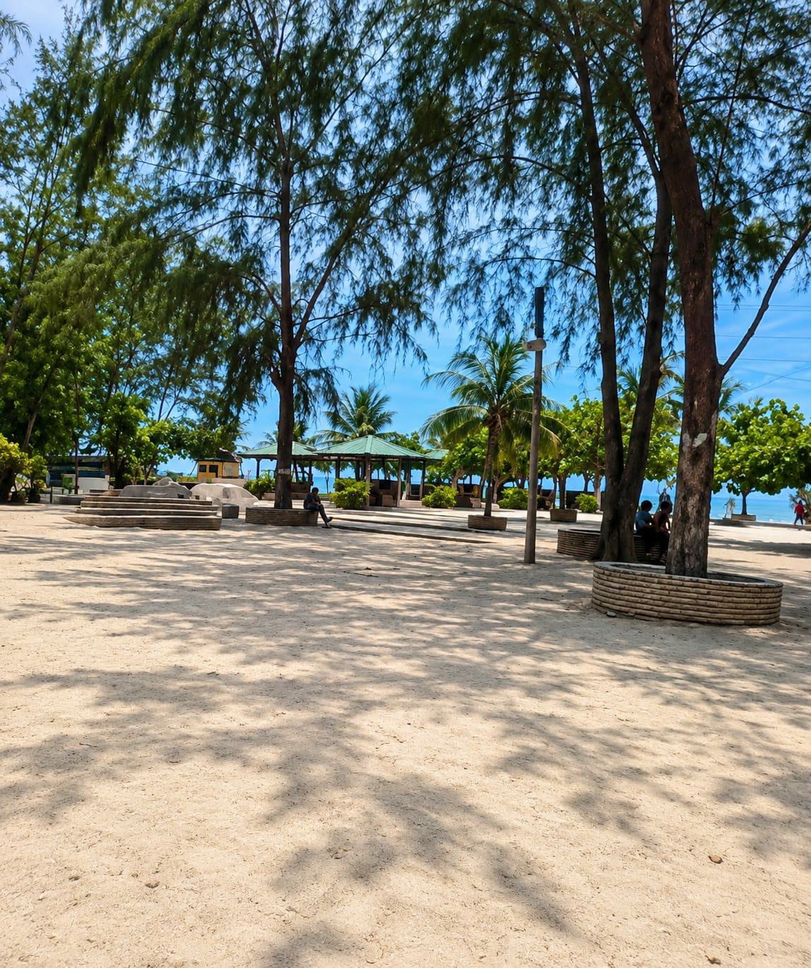 The image depicts a sandy area with scattered trees and palm trees, suggesting a tropical beach or park setting. In the background, there are several open-air pavilions with green roofs, some of which appear to be seating areas. A few individuals are visible: one person is sitting on a stone bench near a tree, another group of people are gathered near a tree, and a couple of individuals are walking in the distance. The ground is covered in sand, with shadows cast by the trees. A concrete structure, possibly a planter or seating area, is visible in the foreground. The sky is blue with some visible clouds. The overall scene suggests a public outdoor recreational space. The location is identified as Kota Pari, Indonesia.
