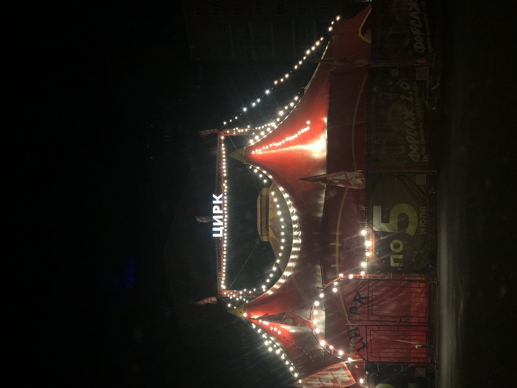 The image shows a nighttime scene of a brightly lit circus tent. The tent is predominantly red with yellow accents and is adorned with strings of warm, white lights that create a festive atmosphere. A prominent sign in Cyrillic letters reads "ЦИРК" (Circus), illuminated by its own set of lights.
Several banners are visible on the side of the tent. One banner displays "ФОРСАЖ 2.0" and below it, "ЦИРК шапито «ЗВЁЗДНЫЙ»" (Circus Tent "Starry"). Another banner, partially visible, also says "ФОРСАЖ". A large "5" is prominently displayed, with the word "Июля" (July) beneath it, indicating a date, likely the start or end of an event. The overall setting is dark, suggesting it is evening or night.
The circus appears to be set up outdoors, with a dark background that could be a field or an open area. The lighting from the circus tent illuminates the immediate surroundings, creating a stark contrast with the darkness. There are no people clearly visible in the image, but the circus tent, lights, and banners strongly suggest an event is either in progress or about to begin. The overall impression is one of excitement and spectacle associated with a circus performance. The location context of Petropavlovsk-Kamchatsky, Russia, adds a specific geographical detail to the scene.